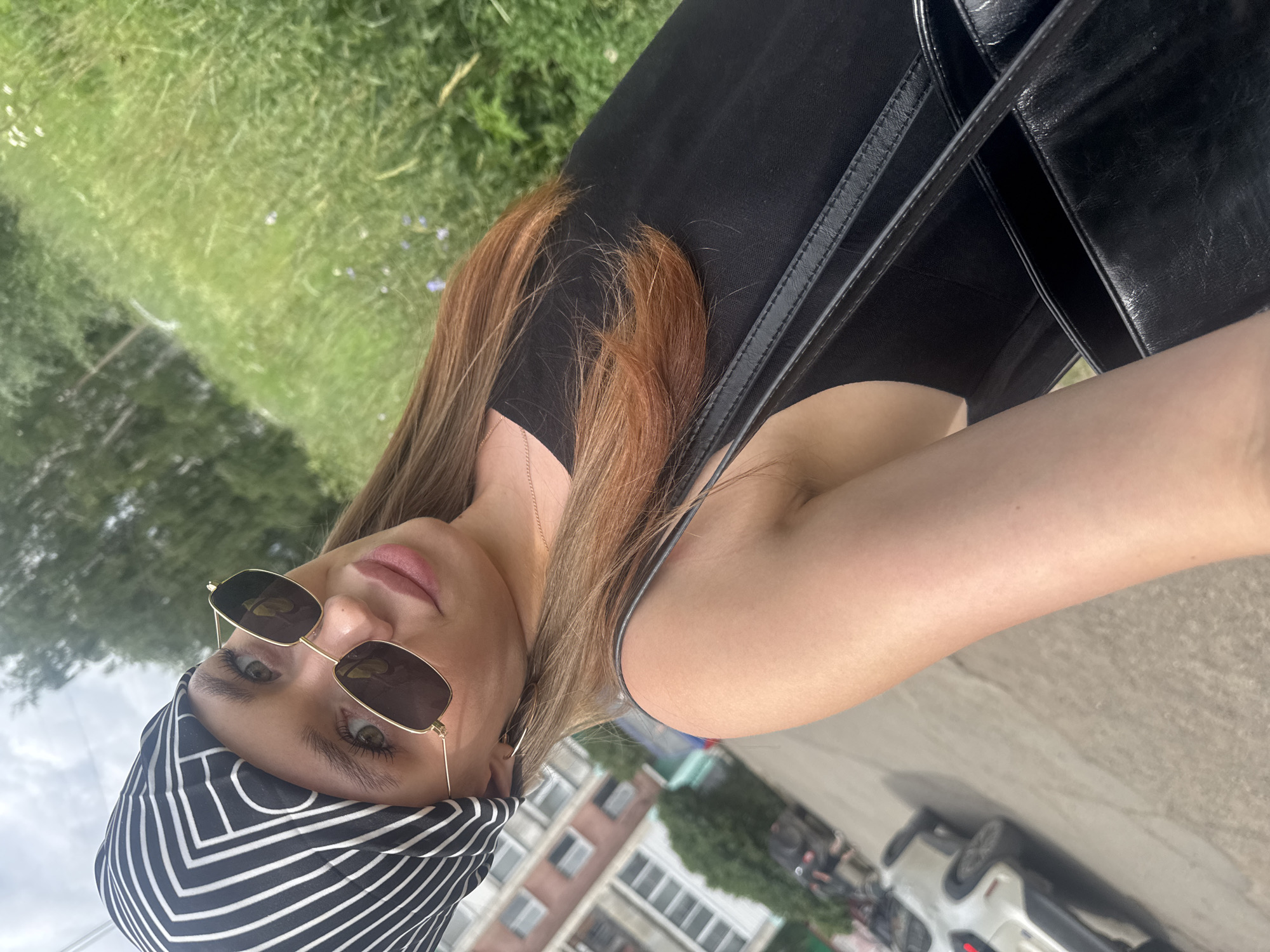 The media file shows a young woman with long, reddish-brown hair, wearing a black top and a black and white striped bandana. She is also wearing gold-rimmed sunglasses. A black purse is visible, slung over her shoulder. The image appears to be a selfie taken outdoors, with a slightly blurred background of green grass and buildings. The woman's face is turned towards the camera, and she has green eyes. The lighting suggests it is daytime, possibly late morning or early afternoon, with bright sunlight. The overall mood seems casual and relaxed, as if she is out for a walk or enjoying a day outdoors. There are no visible signs or text in the image. The background hints at an urban or suburban environment with buildings and a street with cars. The location context of Chaikovsky, Russia, provides a geographical setting for the scene.