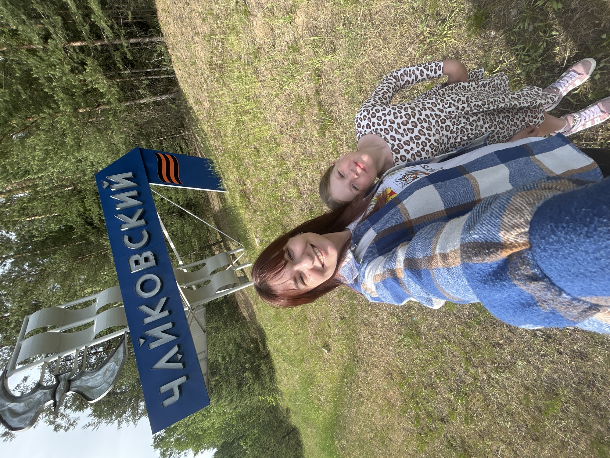 The image shows two people lying on the grass next to a blue sign that reads "Чайковский" (Chaykovsky) in Russian. The sign also features a metallic structure that looks like sails and a bird. One person, likely a woman, is wearing a blue checkered shirt and is smiling at the camera. The second person, likely a young girl, is wearing a leopard-print dress and pink shoes. The background includes a grassy area and trees. The presence of the Chaykovsky sign suggests that the location is in the city of Chaykovsky, Russia.