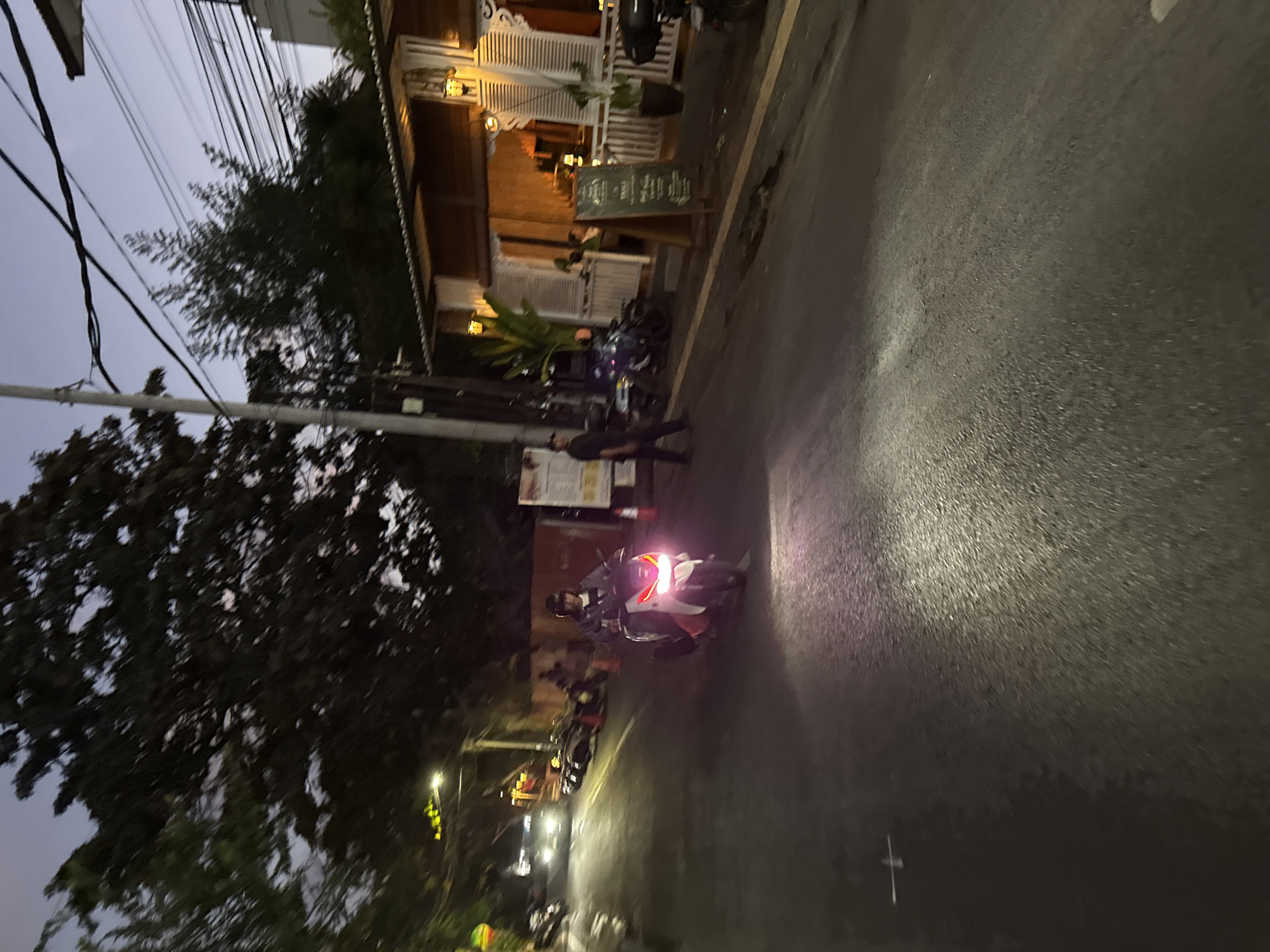 This is a nighttime, eye-level, vertical shot taken on a street in Denpasar, Indonesia, likely near a restaurant or cafe. The sky is a dusky purple, and streetlights and lights from a building cast a warm glow. A person on a white scooter with bright pink headlights is the central focus, illuminated by their own headlight and ambient light. The scooter has a dark seat and rider wearing a helmet and dark clothing. To the right, a wooden building with white shutters and outdoor seating is visible, with a sign that appears to be a menu. A person in dark clothing is walking away from the building and towards the street. In the background, more scooters are parked along the street, and a white car with headlights on is approaching from the left. Trees and power lines are visible against the twilight sky. The road surface is asphalt, wet in places, reflecting the light.