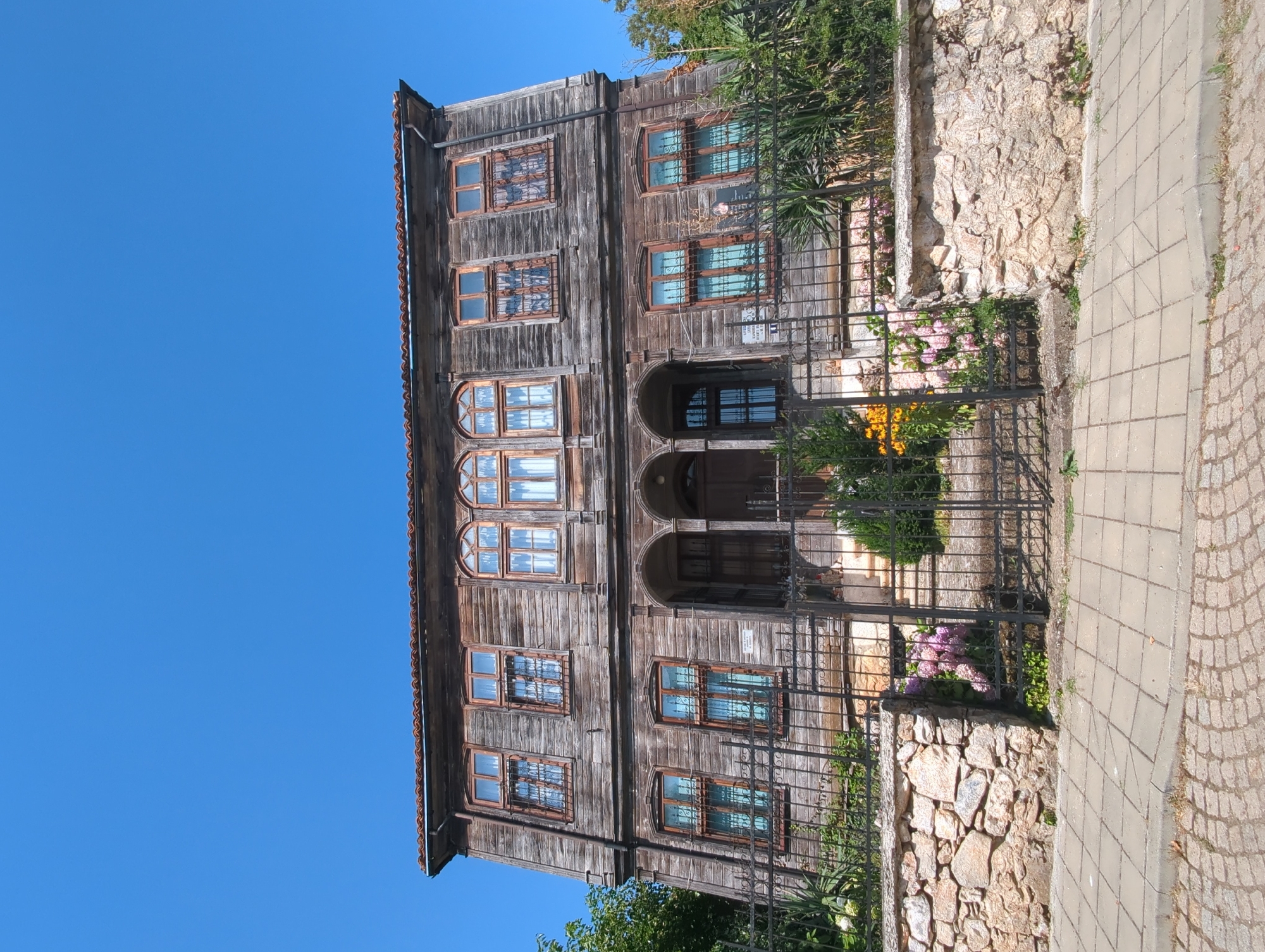 An old, two-story wooden house with a reddish-brown tile roof stands under a clear blue sky. The house is weathered, with visible wood grain and some signs of age. It has numerous windows, many with arched tops, and some have decorative metal grilles. A dark metal fence runs along the front of the property, in front of a stone wall. A paved walkway, made of square concrete slabs, runs along the bottom of the frame, with a cobblestone street visible to the left. Lush green foliage and flowering plants, including hydrangeas and marigolds, are visible in front of the house and behind the fence. The overall scene suggests a historic building in a well-maintained, possibly residential area. This house is located in the city of Malko Tarnovo, Bulgaria.