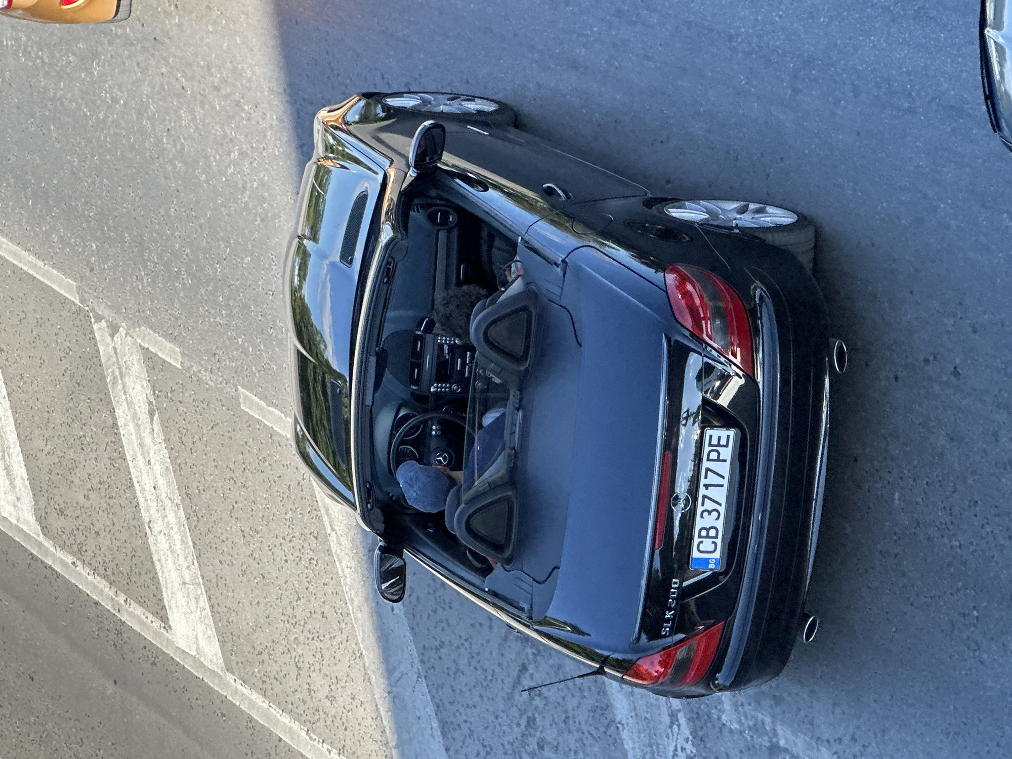 The image shows a black Mercedes-Benz SLK 200 convertible driving on a road in Veliko Tarnovo, Bulgaria. The license plate reads CB 3717 PE. There are two people in the car. The driver, wearing a blue baseball cap, is visible from the back. A person with dark, curly hair is in the passenger seat, also viewed from the back. The car is on a paved road with white lane markings. The scene appears to be a street in a city.