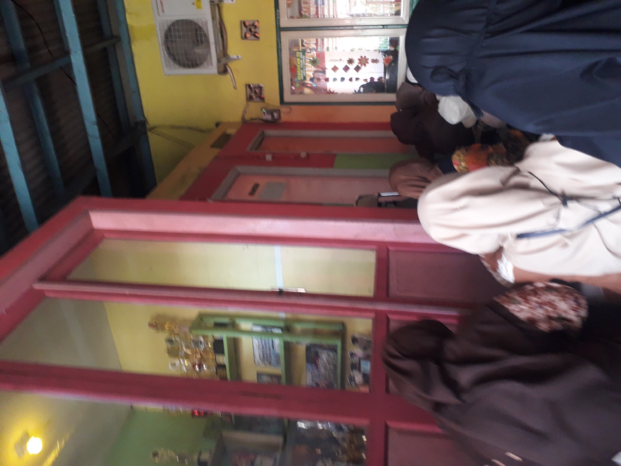 In Cankuang, Indonesia, an indoor scene depicts several people, predominantly women, wearing headscarves and facing away from the camera. They appear to be seated or kneeling in a gathering. The environment features brightly colored walls, with a yellow wall visible on the right and a greenish wall on the left. A red-framed doorway or partition dominates the foreground. Behind the people, a shelf displays trophies and framed items. An air conditioning unit is mounted on the yellow wall. The flooring is not clearly visible. The lighting appears to be artificial, with a bright light fixture visible in the upper left corner.