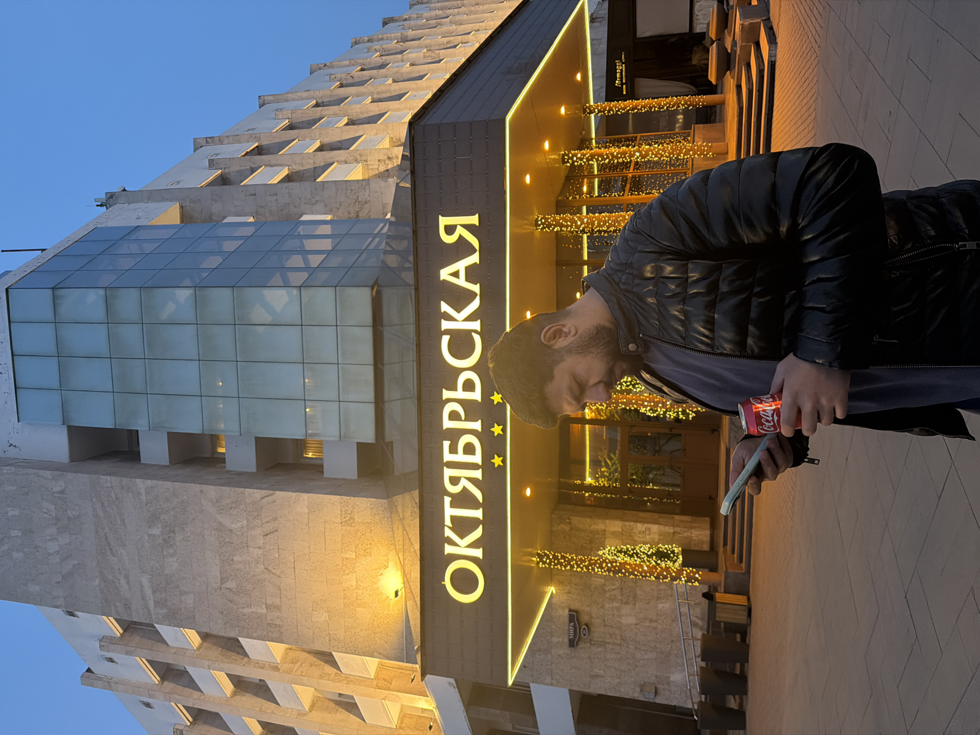 A person stands outside the Oktyabrskaya Hotel in Krasnoyarsk, Russia. The individual holds a mobile phone in their left hand and a can of Coca-Cola in their right hand. The hotel's facade, with "ОКТЯБРЬСКАЯ" (Oktyabrskaya) lit up in yellow, is visible in the background, along with festive string lights. The building architecture is visible, including a modern glass structure atop a stone base. The sky is a pale blue, indicating evening or early morning light.