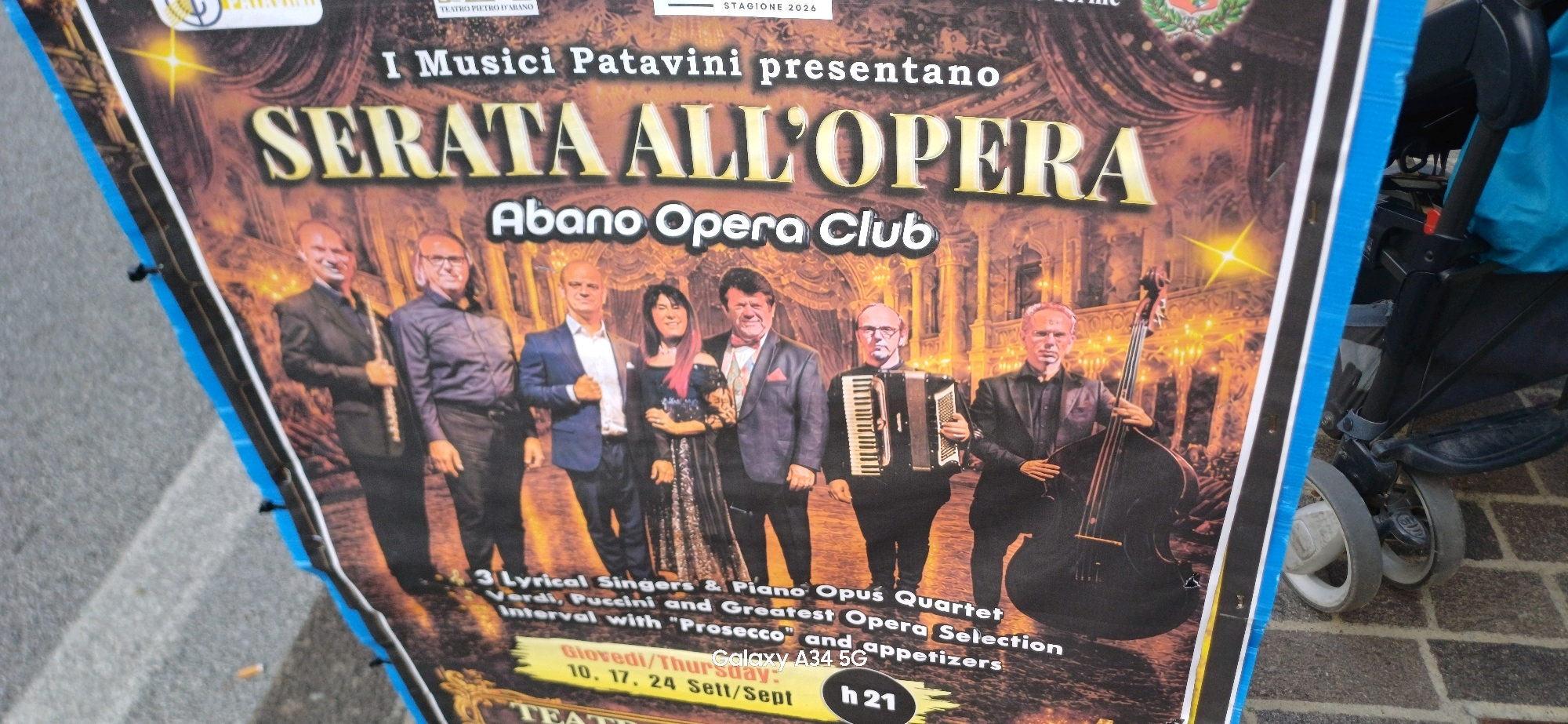 The image is a photograph of a poster advertising an opera event in Abano Terme, Italy. The poster features an image of a musical ensemble, including a man playing a double bass, another playing an accordion, and several singers. The text on the poster reads "I Musici Patavini presentano SERATA ALL'OPERA Abano Opera Club" and lists the dates "10, 17, 24 Sett/Sept" and the start time "h 21". The poster is outdoors, with a portion of a stroller visible on the right side of the image. The ground appears to be paved with cobblestones.