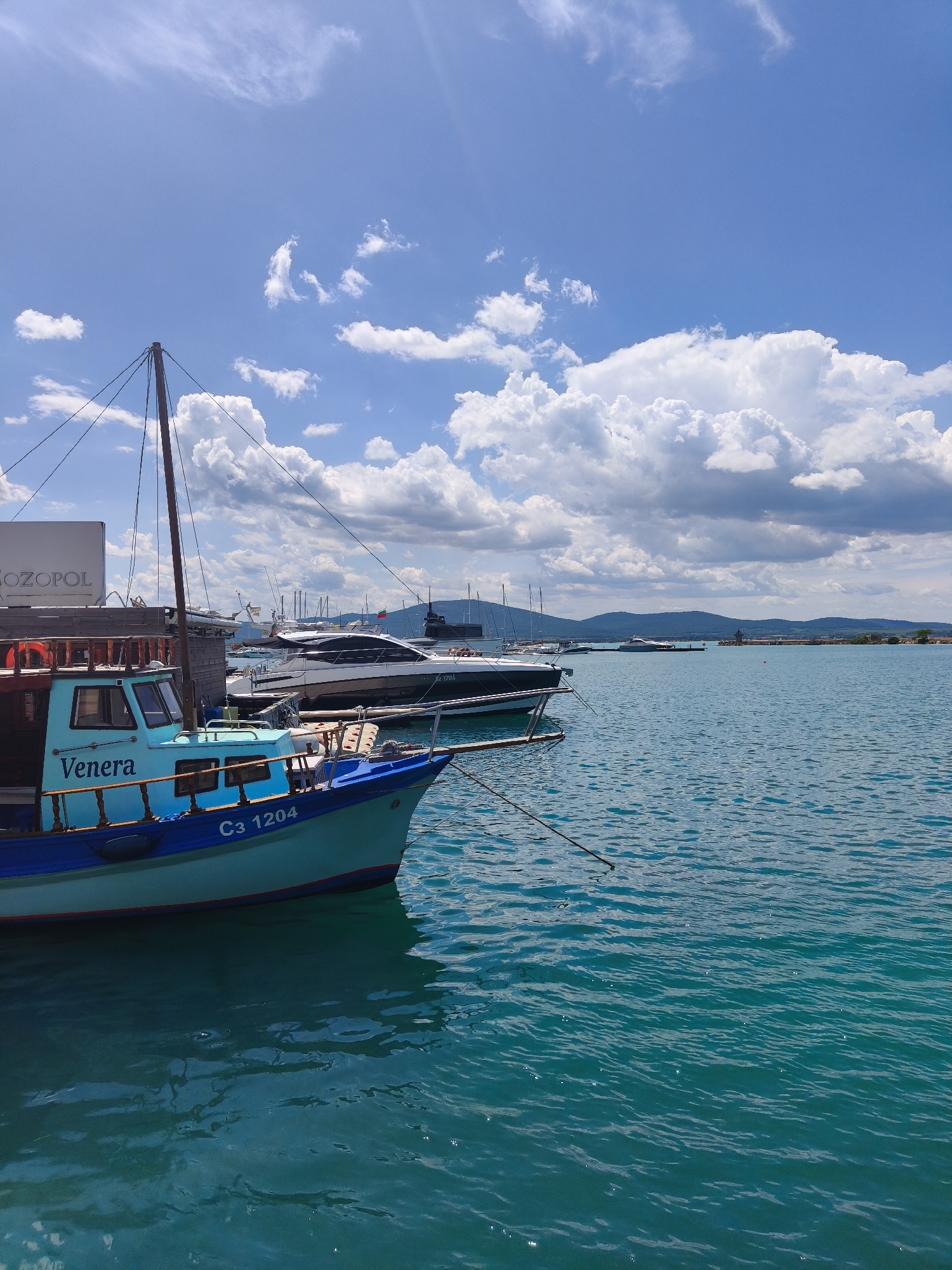 The image presents a sunny daytime scene in Sozopol, Bulgaria. The foreground features a light blue and dark blue fishing boat named "Venera" with Cyrillic letters reading "C3 1204" on its hull. The boat is moored in turquoise water, and a rope extends from its bow. Behind the "Venera," several yachts are docked. In the background, there is a visible land mass with a coastal appearance and some buildings and mountain range on the distant horizon. The sky is bright blue with scattered cumulus clouds. A building with the word "Sozopol" on it is partially visible on the left.