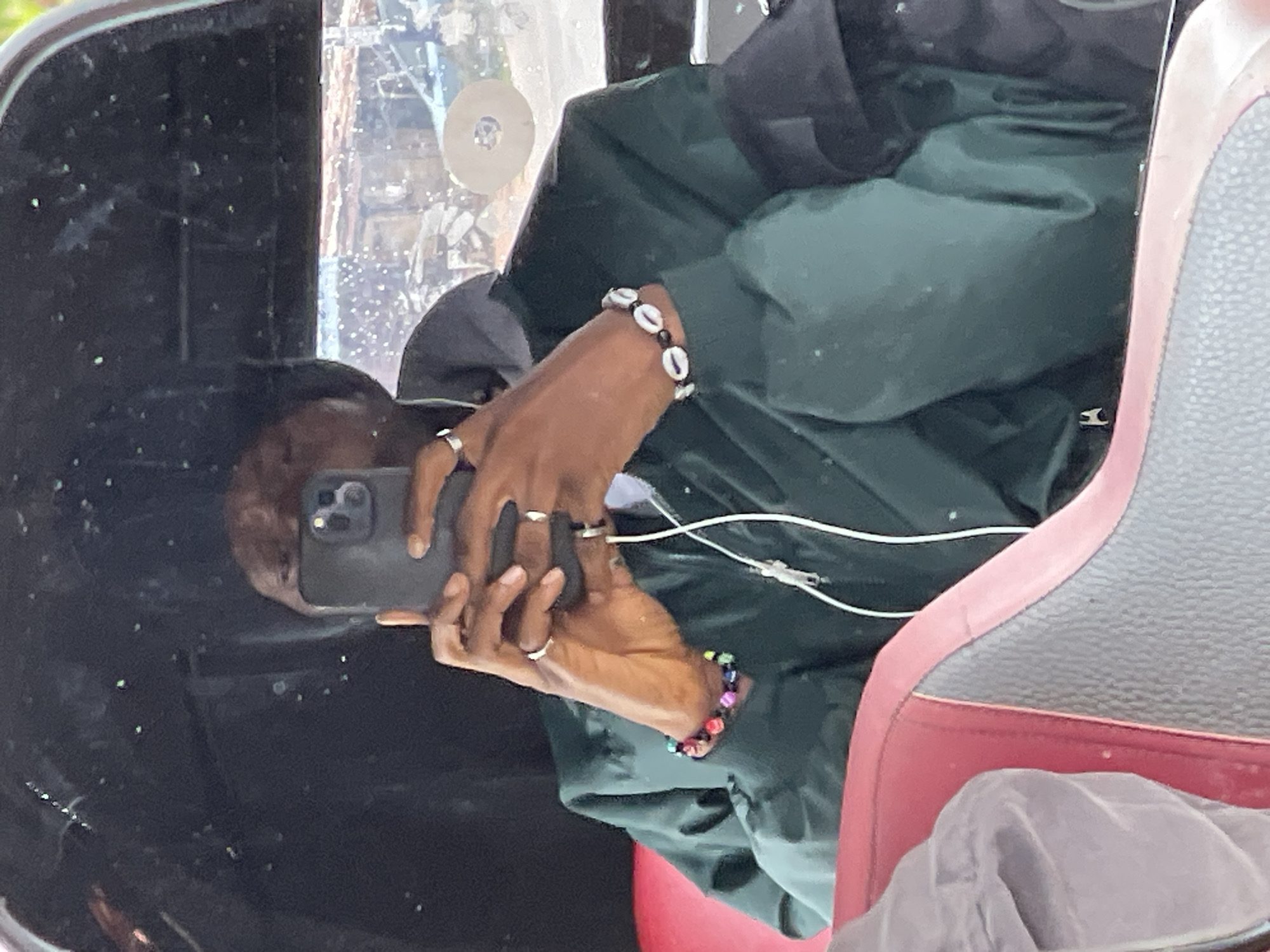 The image, rotated 90 degrees counter-clockwise, captures a person inside what appears to be a vehicle. The individual, with dark skin, is seen from the chest up, holding a black smartphone, possibly an iPhone, with both hands. Their face is largely obscured by the device, suggesting they might be taking a photo, recording a video, or viewing the screen.
The person is dressed in a dark green jacket or hoodie, with a black cap or hood visible on their head. Their hands are adorned with several silver-toned rings. They also wear two bracelets: one on the left wrist featuring white cowrie shells alternating with black beads, and another on the right wrist made of vibrant, multi-colored beads. A white wired earbud cable is visible, extending from the phone.
The setting appears to be the interior of a vehicle, possibly a car or bus, with a section of a red and grey patterned seat visible on the right. A window occupies the upper and left parts of the frame, through which a blurred outdoor environment can be seen, indicating daytime. The window surface shows specks of dirt or dust, along with reflections.
The scene takes place in Jos, Nigeria. No visible text is present in the image.