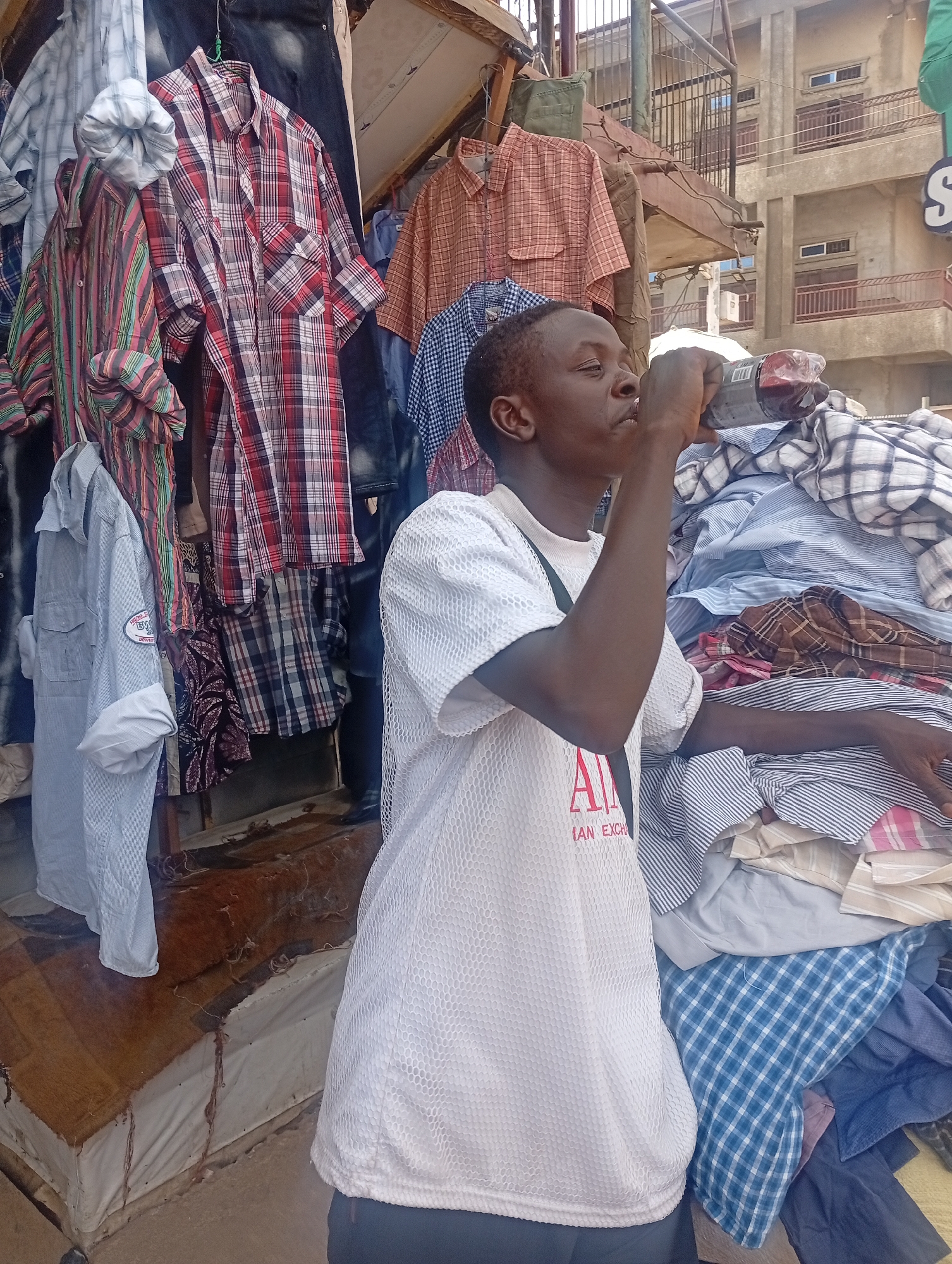 A young man is drinking from a plastic bottle in front of a stall selling used clothes in a market in what is purported to be Maiburiji, Nigeria. He is wearing a white t-shirt with a logo and black trousers. The stall displays a variety of button-down shirts in various colors and patterns, hung on hangers or piled in stacks. The background shows a multi-story building and a metal structure, suggesting an urban commercial environment.