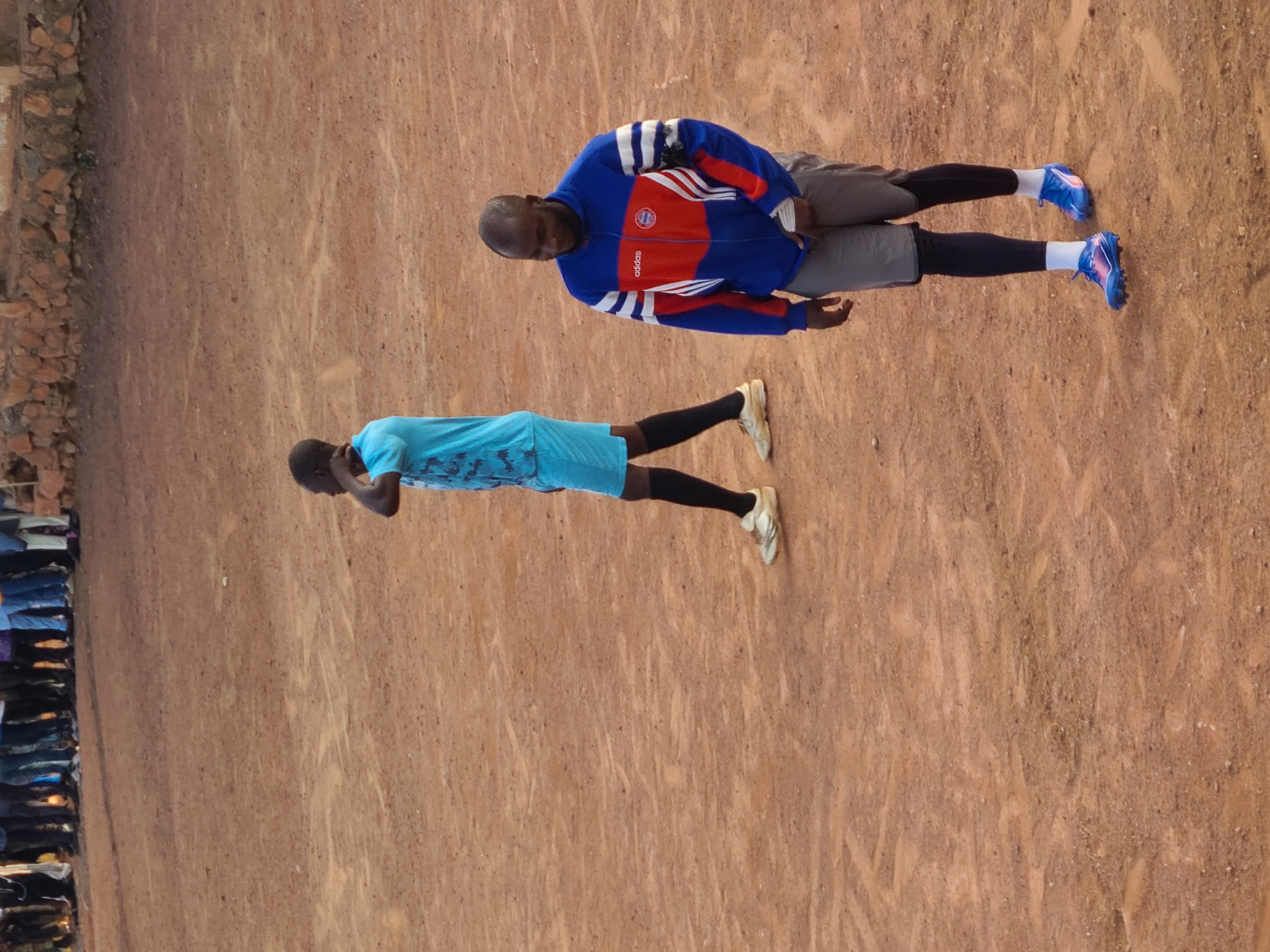 This image depicts an unpaved, reddish-brown earth surface, likely a sports field, under natural light. In the foreground, positioned right of center, stands a dark-skinned adult male. He wears a blue, bright red, and white full-zip track jacket with "adidas" printed in white on the red chest section, and a circular emblem below it. White stripes adorn the sleeves. He has grey athletic shorts over black compression leggings, white socks, and blue cleated athletic shoes with a pink/purple accent. His head is bald or closely shaven, and he has a short beard. He looks downward and slightly to his left, with his right hand partially obscured in front of him.
Further back and to the left, a second dark-skinned adult male is visible. He wears a light blue short-sleeved athletic shirt with a subtle darker blue camouflage-like pattern, and matching light blue athletic shorts. He has black calf-length socks and off-white cleated athletic shoes. His hair is very short. He is facing slightly to his left, with his head turned to his right, and his right hand is raised to his neck or ear, possibly stretching or adjusting something.
In the blurred background, a line of indistinct figures, likely spectators or other participants, stands along a boundary. Above them, a low wall constructed of reddish-brown stones and earth is visible, topped by a lighter-colored building or wall structure on the far right. The ground is dry and appears to have slight undulations and faint marks from activity. There are no direct interactions between the two prominent figures.
