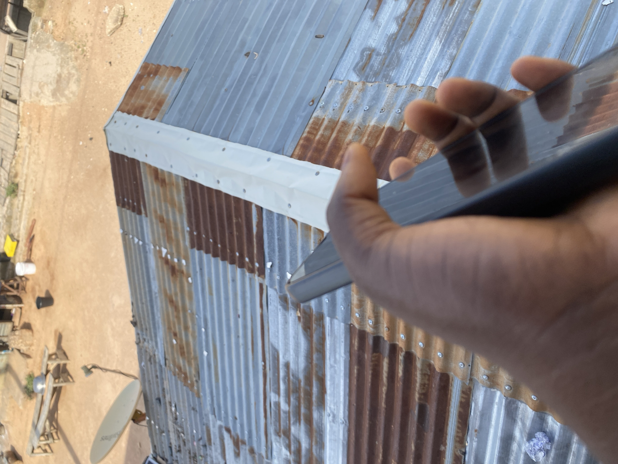 An over-the-shoulder shot shows a dark-skinned person's hand holding a dark smartphone, pointed towards a corrugated metal roof. The roof appears old and rusted in places, with visible seams and fasteners. In the background, out of focus, is an outdoor area with a wooden bench, a satellite dish, and some structures, suggesting a residential or informal settlement. The environment is characterized by dry, dusty ground. This scene likely depicts an informal settlement in Jalingo, Nigeria, given the architectural style of the roof and the general appearance of the surroundings.