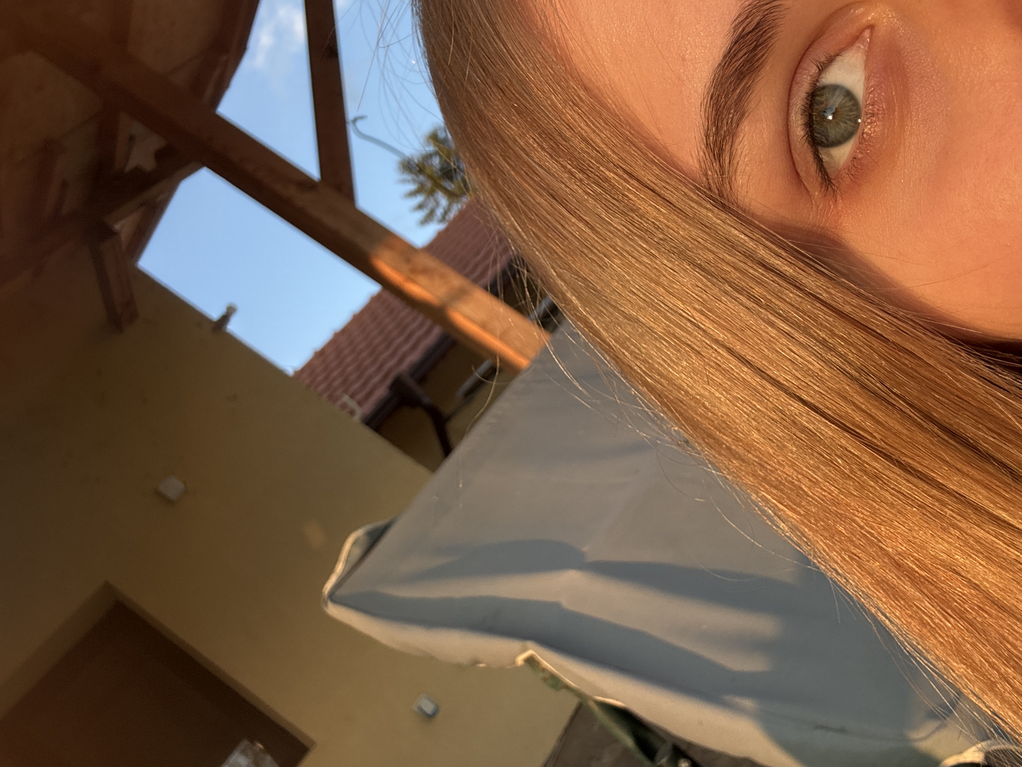 The image shows a close-up view of a person's face, specifically the right eye and eyebrow, with long blonde hair partially covering the face. The eye is green with visible details of the iris. The background includes a blurred outdoor scene. An architectural structure with wooden beams is visible against a blue sky. A building with a red tile roof is also present. A white, possibly fabric-covered object is in the lower portion of the background. The scene appears to be outdoors, likely taken during the day with natural sunlight illuminating the subject. There is no specific indication of the location being in Lehchevo, Bulgaria, in the image.