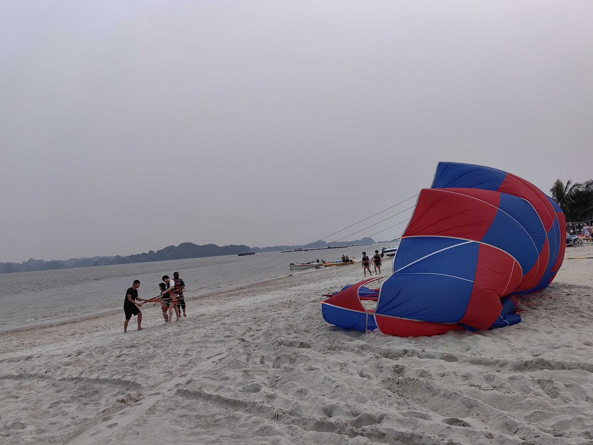The image depicts a beach scene in Bai Chay Ward, Vietnam, under an overcast sky. In the foreground, a large, deflated parasailing parachute lies on the white sand. The parachute features a pattern of alternating red and blue panels. Several people are standing on the beach, some holding onto ropes connected to the parachute. There appear to be four people towards the left, pulling on the ropes. Other individuals are walking near small boats on the water's edge in the distance. The water is calm, and there are landmasses or islands visible on the horizon. In the background to the right are beach trees and a few people on the shore. The sand shows numerous footprints and drag marks.