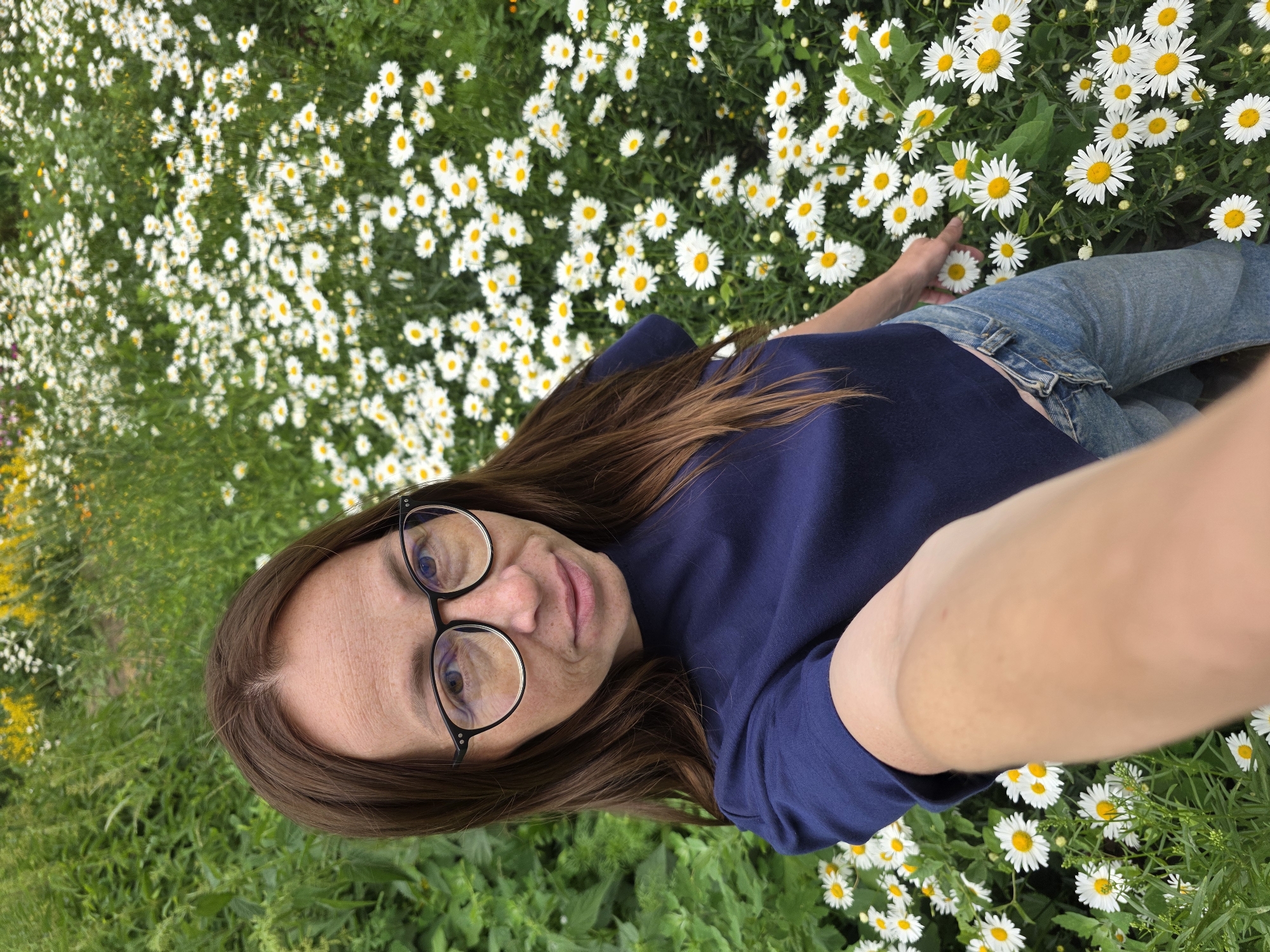 The image shows a woman with brown hair, wearing glasses, a navy blue t-shirt, and blue jeans, standing in a field of white daisies. She is holding her phone up to take a selfie. The field of daisies is dense, with some yellow flowers mixed in. The background is blurred, but appears to be more greenery and flowers. The location is Chaikovsky, Russia.