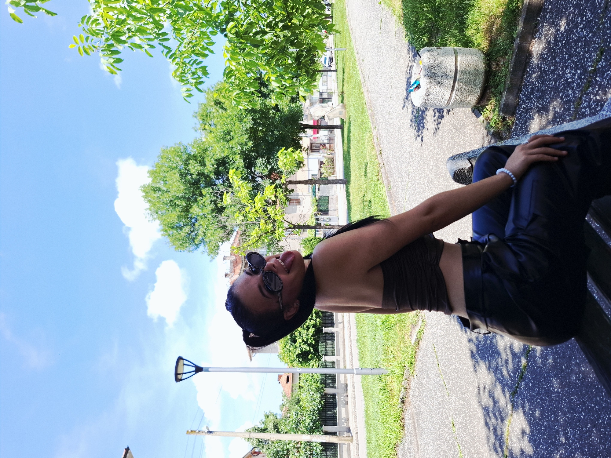 The image shows a young woman sitting on a park bench outdoors in Mezdra, Bulgaria, under a clear, sunny sky with some white clouds. She is positioned on the right side of the frame, looking playfully towards the camera with her tongue slightly extended. She wears dark sunglasses, a dark brown, strapless-style top, and dark, possibly leather, trousers. Her dark hair is pulled back, and a light-colored beaded bracelet is visible on her right wrist, which rests on her lap.
The setting appears to be a public green space or park with a paved path. Lush green trees are abundant, casting shadows on the ground, indicating bright daylight. In the background, beyond the trees, several buildings are visible, suggesting an urban or suburban environment. A modern-style street light is seen against the sky on the left side of the frame. Near the bench, a light-colored, cylindrical concrete object, possibly a planter or bollard, is also present. There is no visible text in the image. The overall impression is one of leisure and cheerfulness on a bright day.