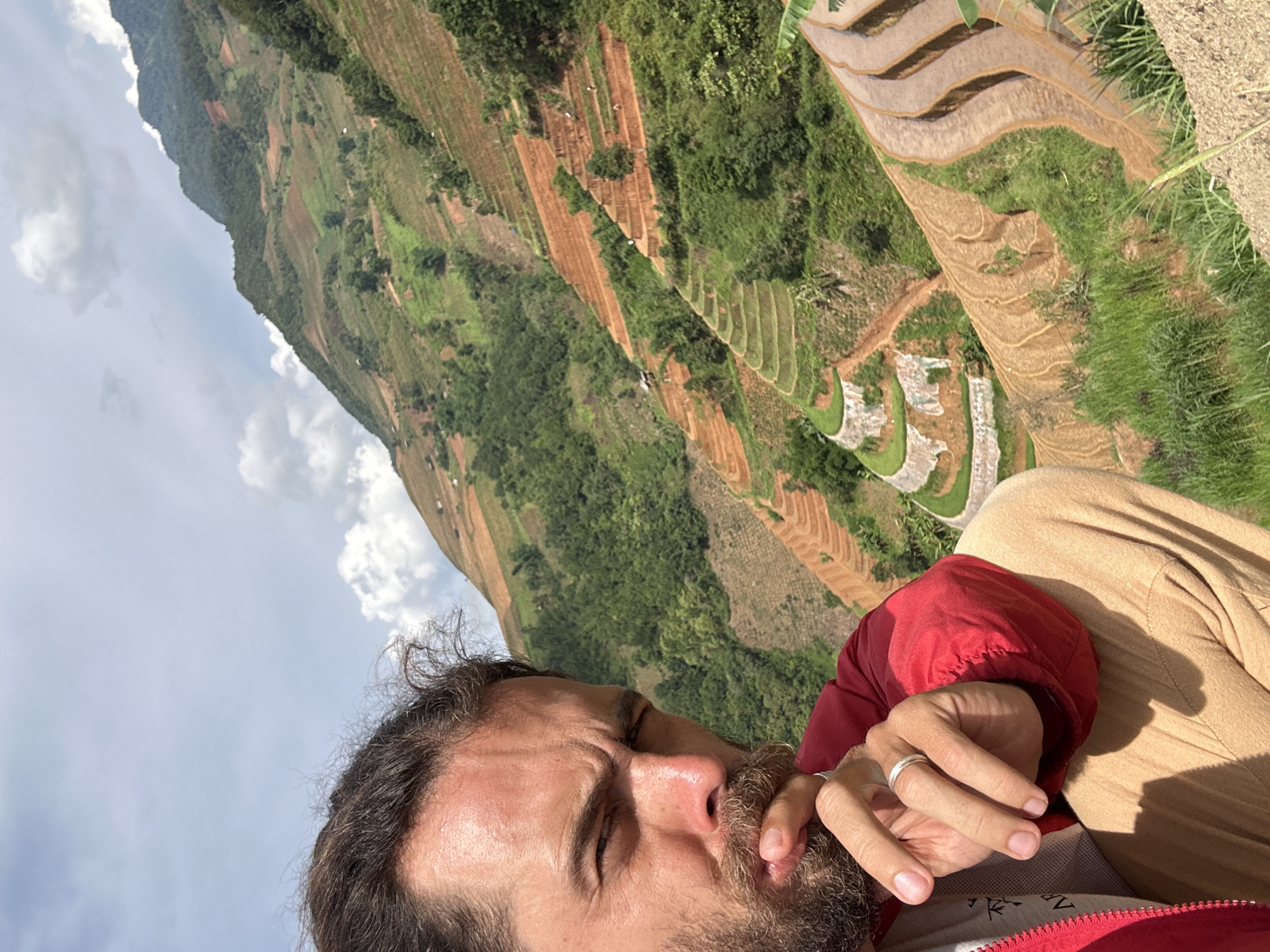 The image displays a rotated portrait of a person in the foreground, set against a backdrop of mountainous terraced fields under a bright, cloudy sky. The image is rotated 90 degrees clockwise, placing the horizon vertically on the left side of the frame.
In the lower right foreground, a man is partially visible. He has dark, curly hair and a full beard. His eyes are squinted or closed, possibly due to direct sunlight. His right hand is raised towards his face, with a silver ring clearly visible on his ring finger. He is wearing a red jacket, part of a grey shirt is visible underneath, and light brown or beige pants.
The background reveals a spectacular landscape of terraced fields, indicative of rice paddies. These terraces are carved into steep mountain slopes, displaying alternating patterns of reddish-brown soil and vibrant green vegetation. Further up the slopes, denser green foliage covers the mountains. The sky, filled with scattered white clouds, dominates the left side of the frame.
The scene depicts a moment of rest or contemplation, with the man seemingly taking in the expansive view of the terraced mountains. The strong, clear daylight with visible shadows suggests it is daytime. The distinctive terraced landscape points to a location like Lao Chai, Vietnam. There is no visible text in the image.