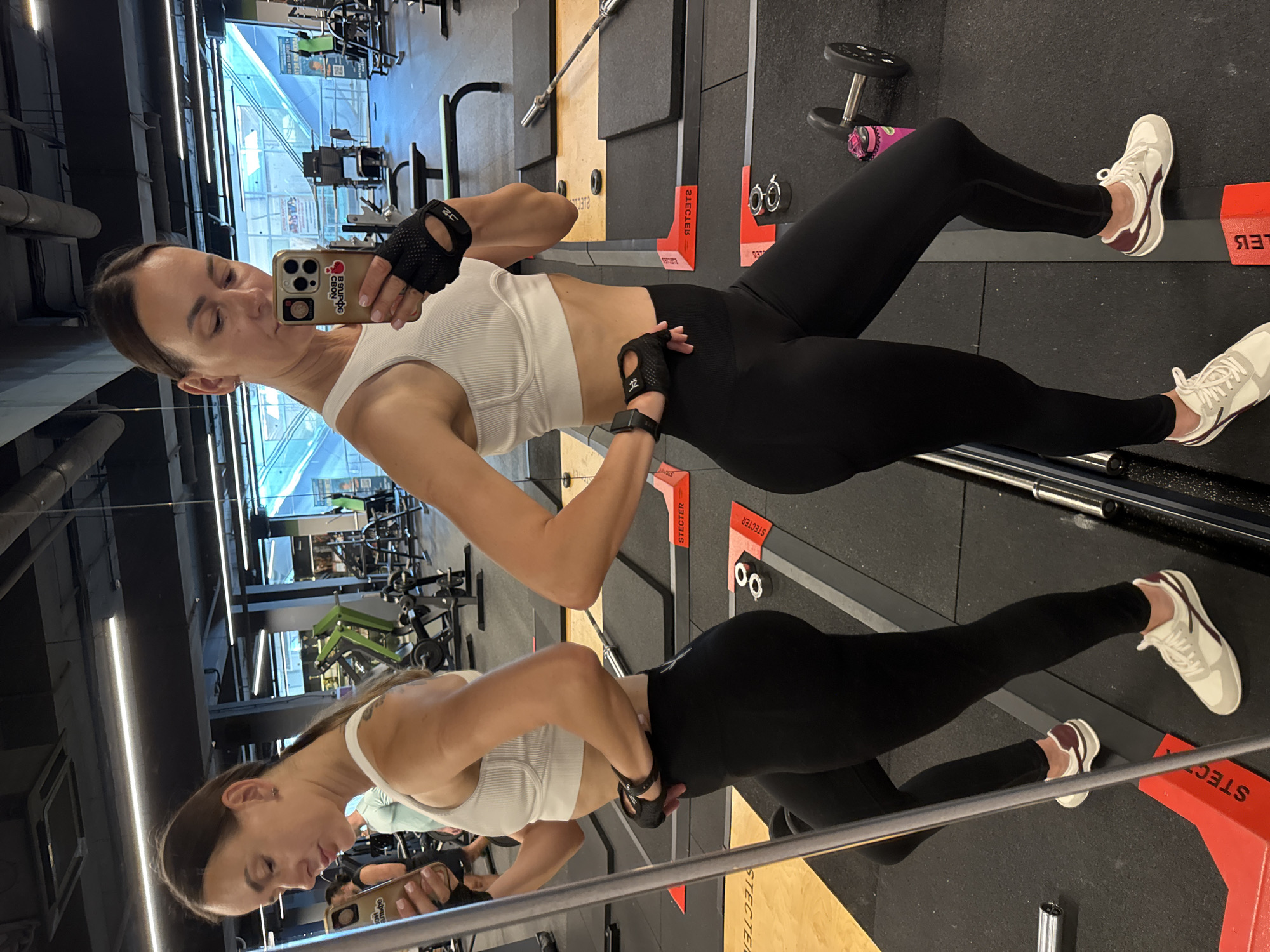 The image shows a woman taking a mirror selfie in a gym. She is wearing a white sports bra, black leggings, and white athletic shoes. She has on black workout gloves. Her hair is pulled back in a ponytail. In the background, there are various gym equipment, including weight racks, benches, and dumbbells. The gym appears to be modern and well-equipped. The location is stated to be in the city of Sochi, Russia.