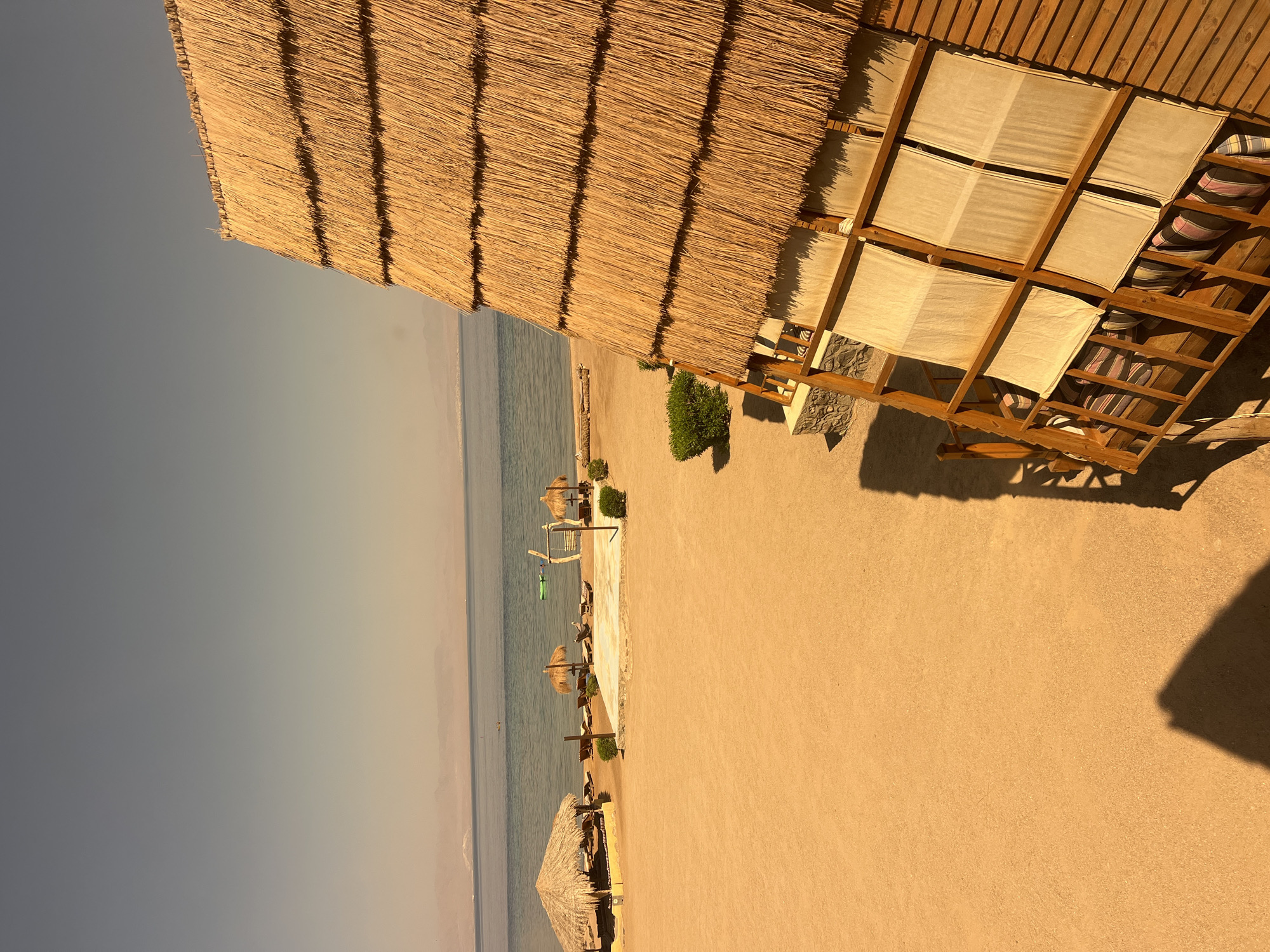 The image depicts a beachfront scene in Nuweiba, Egypt, characterized by a sandy shore, calm turquoise water, and a clear sky. In the foreground, the thatched roof and wooden structure of a beachfront dwelling or cabana are prominently featured. The dwelling has multiple windows covered with light-colored fabric shades. The sandy beach is sparsely populated with lounge chairs and straw umbrellas. In the middle ground, a larger thatched-roof structure provides shade, and a swing set is visible near the water's edge. The water is still, reflecting the muted colors of the sky and distant hazy mountains. The overall atmosphere is tranquil and suggestive of a relaxed resort environment.