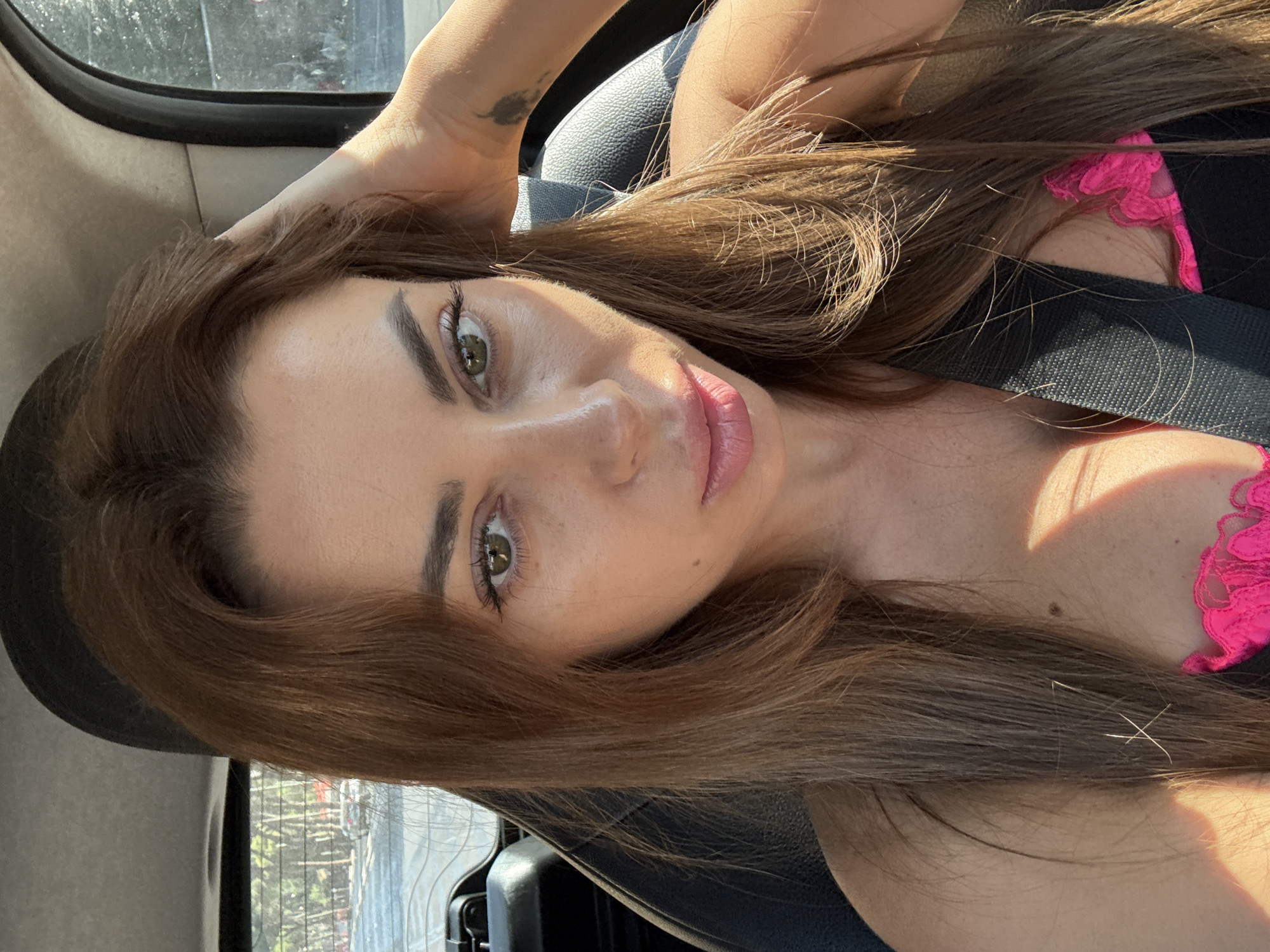 A woman is taking a selfie inside a car. She has long, brown hair and green eyes. She is wearing a pink and black top. The car appears to be parked on a street with trees and buildings visible outside the windows. The sunlight is casting shadows on her face.