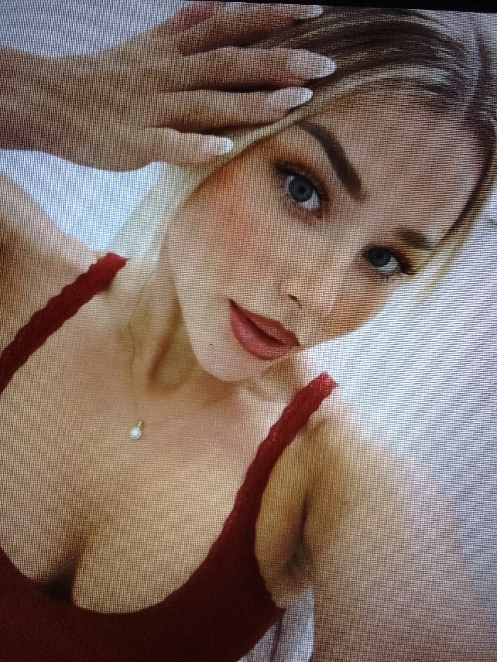 A female individual is depicted from the chest up, positioned at an angle with her head tilted. She has blonde hair and blue eyes, gazing directly at the viewer. Her face shows applied makeup, including eyeshadow, blush, and reddish-pink lipstick. She is wearing a red spaghetti-strap garment. A small necklace with a circular clear stone pendant is visible on her sternum. Her right hand is raised, with manicured fingers (long, light-colored nails) positioned against her temple area. The background is a plain, light-colored surface, indicating an indoor setting. The location is Unknown, Unknown.