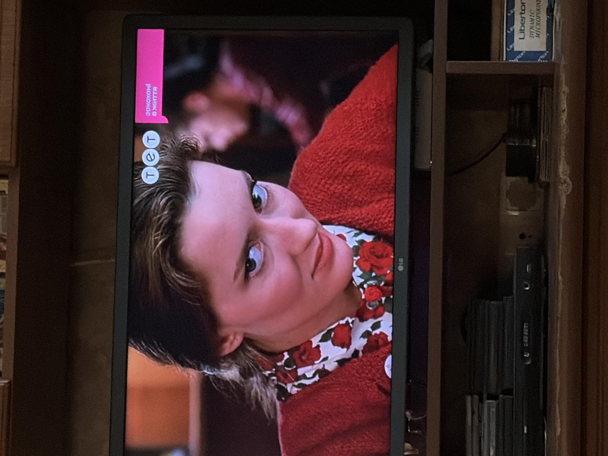The image captures a medium shot of a television screen displaying a scene from a program. On the screen, a woman with brown hair, wearing a red knitted cardigan over a white shirt with a floral print, is looking towards the viewer. She has a direct and somewhat intense gaze. To the upper right of the screen, a pink graphic with white text reads "закохані в життя" and a logo with three circles containing the letters "т е т". Below the television, a shelf is visible, holding a stack of DVDs or similar cases and a DVD player. To the right of the DVD player, a blue and white box labeled "Liberton DYNAMIC MICROPHONE" is present. The background appears to be a dimly lit room with wooden shelving. The television itself is mounted on a stand. The scene is indoors, and the overall impression is that of a home entertainment setup.