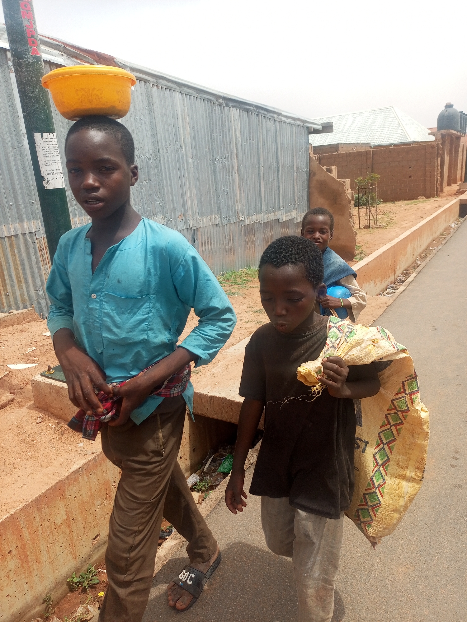 Three boys are walking along a paved road in Tempure, Nigeria. The boy in the front has a yellow bowl on his head, is wearing a turquoise shirt, and brown pants with sandals. The boy next to him is carrying a large yellow sack. A third boy, holding a blue bowl or ball, is walking behind them. The setting is a neighborhood with a corrugated metal building, a red brick structure, and a water tank. The road is flanked by a concrete curb and sandy dirt.