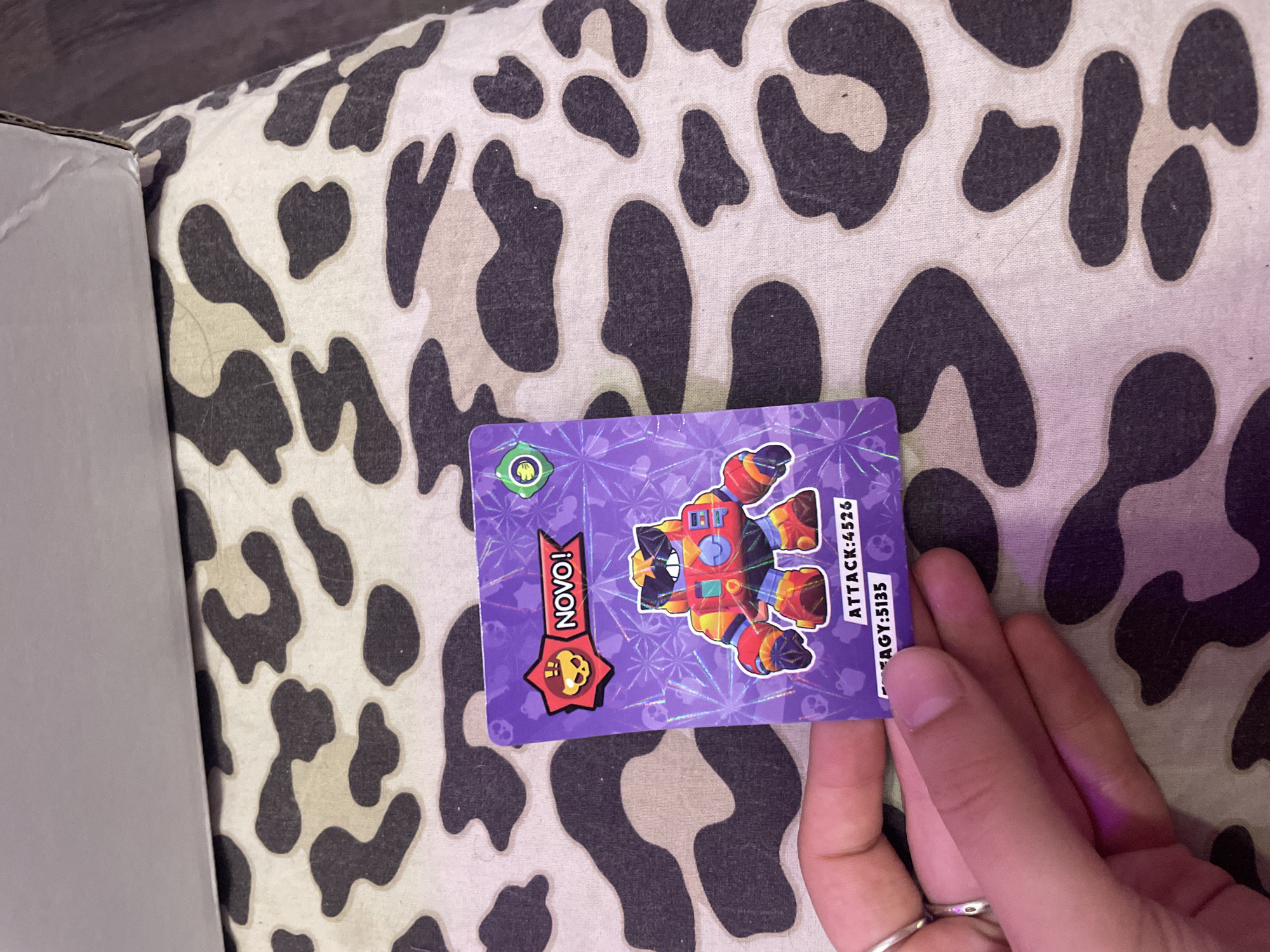 A person's hand, adorned with two silver rings on the index and middle fingers, holds up a purple trading card featuring a cartoon character with sunglasses and a red, orange, and yellow robotic body. The card is positioned over a patterned fabric with brown and beige splotches resembling a cow print. Text on the card reads "NOVO!" and "ATTACK: 4526". The background is out of focus, but a corner of a white cardboard box is visible in the upper portion of the image. The scene appears to be indoors, with artificial lighting casting a purple hue on the hand. The location is not identifiable as Vaganovo, Russia, or any specific city.