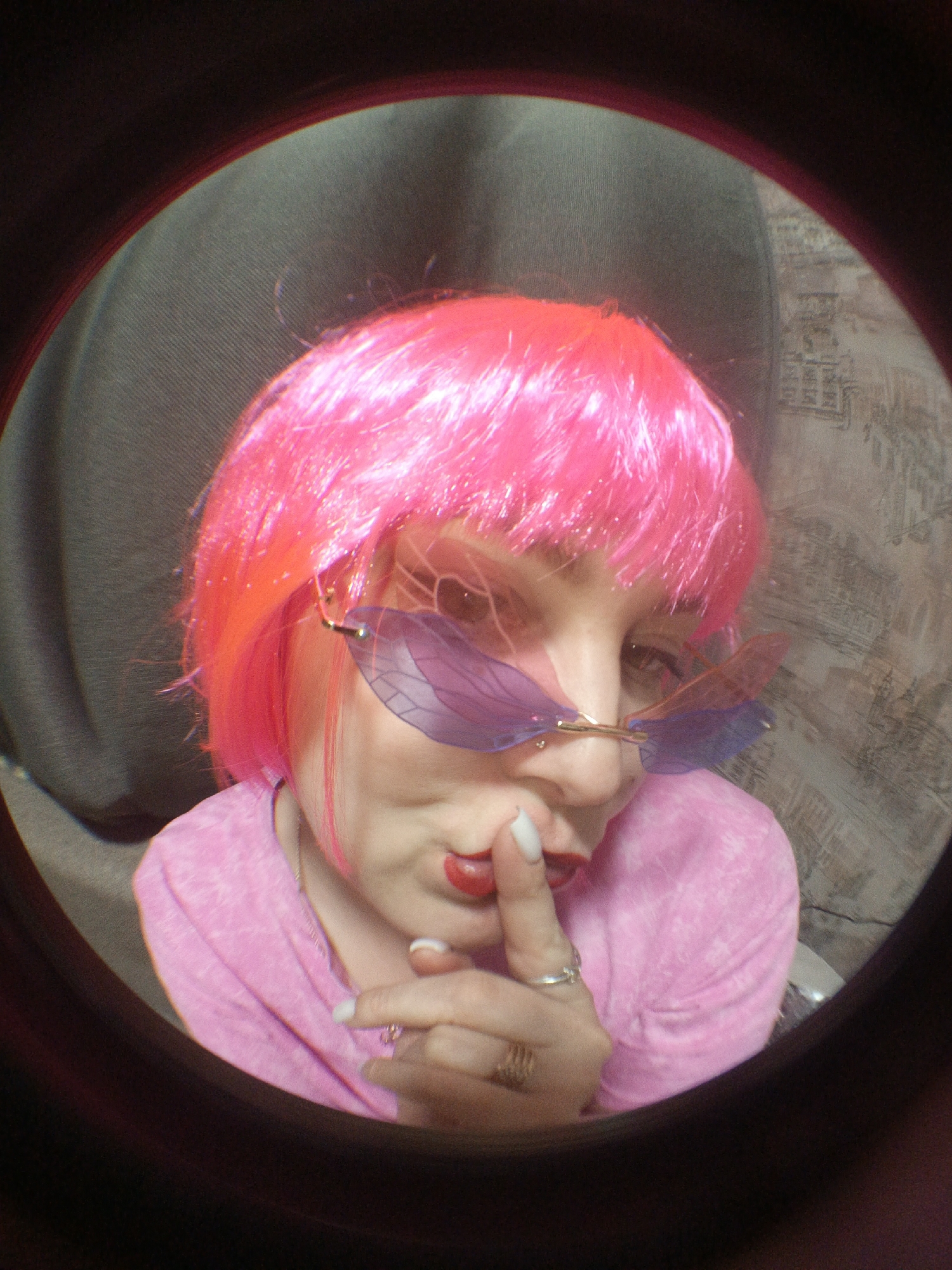 A person with pink hair, wearing sunglasses that resemble dragonfly wings, is holding a finger to their lips. They are wearing a pink shirt. The background shows a gray curtain and patterned wall. The image is framed by a black circle, suggesting it was taken through a lens. The location is Марковский, Russia.