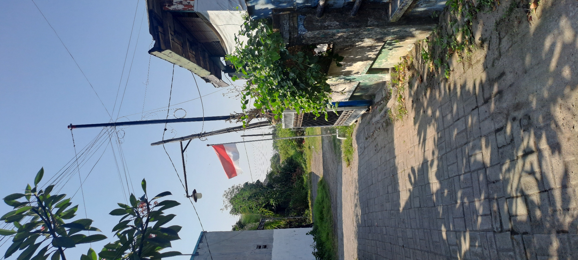 The image shows a street scene in Cemandi, Indonesia. A paved walkway leads away from the viewer, with a shadow of leaves cast on it. To the left, a white building stands with a blue trim. To the right, a wall painted with green and blue accents borders the walkway. A metal gate is partially open, revealing a dense patch of green foliage behind it. A flag, red and white, is mounted on a pole in the mid-ground, waving in the breeze. A lamppost is also visible to the left of the flag. Overhead, a tangle of electrical wires crisscrosses the clear blue sky. In the background, a dirt path curves through greenery. The overall impression is of a sunny day in a semi-urban or rural residential area.