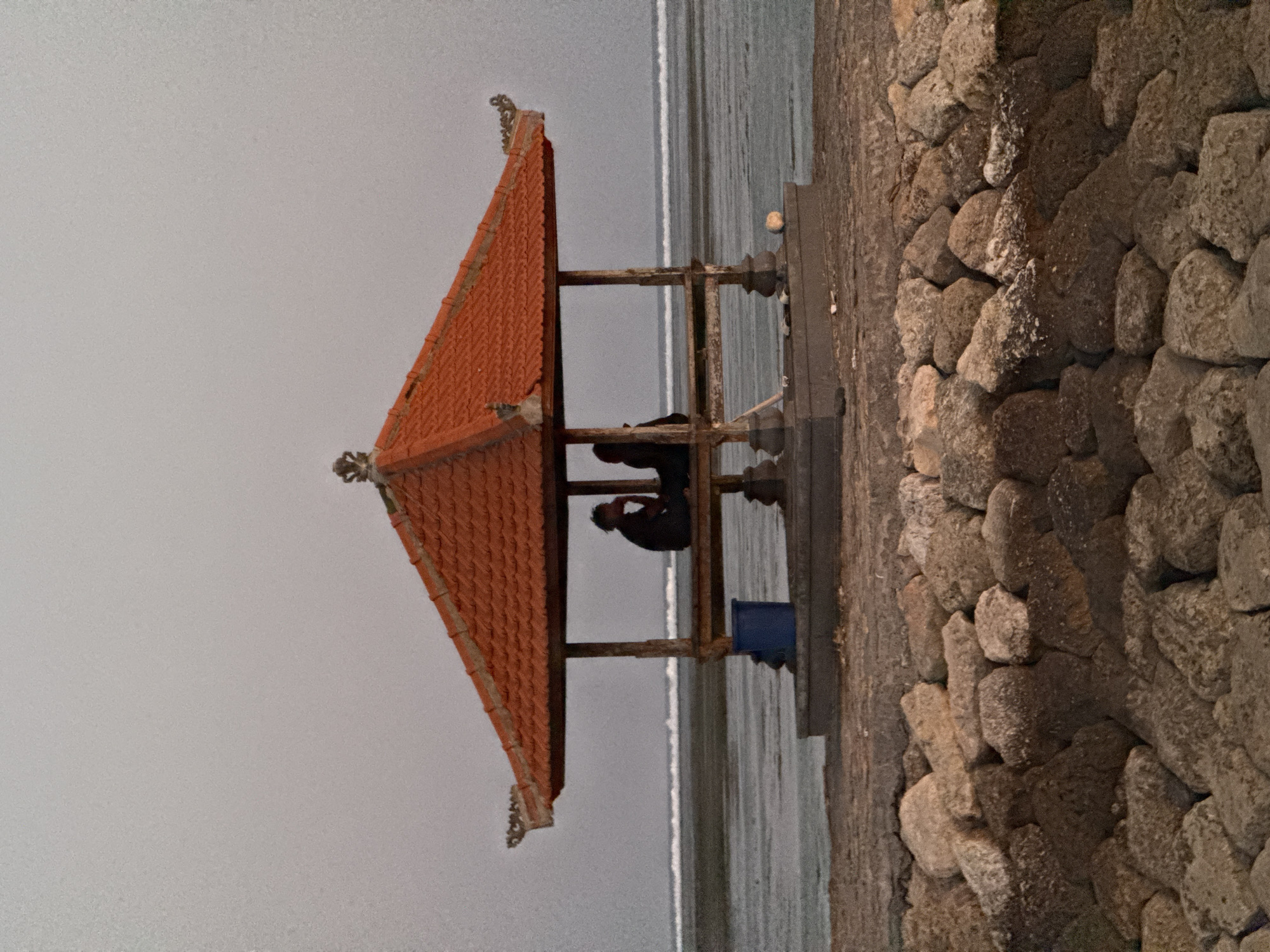 In Denpasar, Indonesia, a gazebo with a distinctive orange tiled roof stands on a concrete platform overlooking the ocean. Two individuals are seated within the gazebo, their figures silhouetted against the water. One person is facing left, leaning forward with a hand near their face. The other person is facing right, also seated. A blue bucket is visible to the left of the gazebo, and a light-colored object, possibly a ball, rests on the platform to the right. The foreground is a jumble of large, textured rocks. The ocean stretches out behind the gazebo, with a distinct line of waves breaking near the shore. The sky is overcast and a neutral gray.