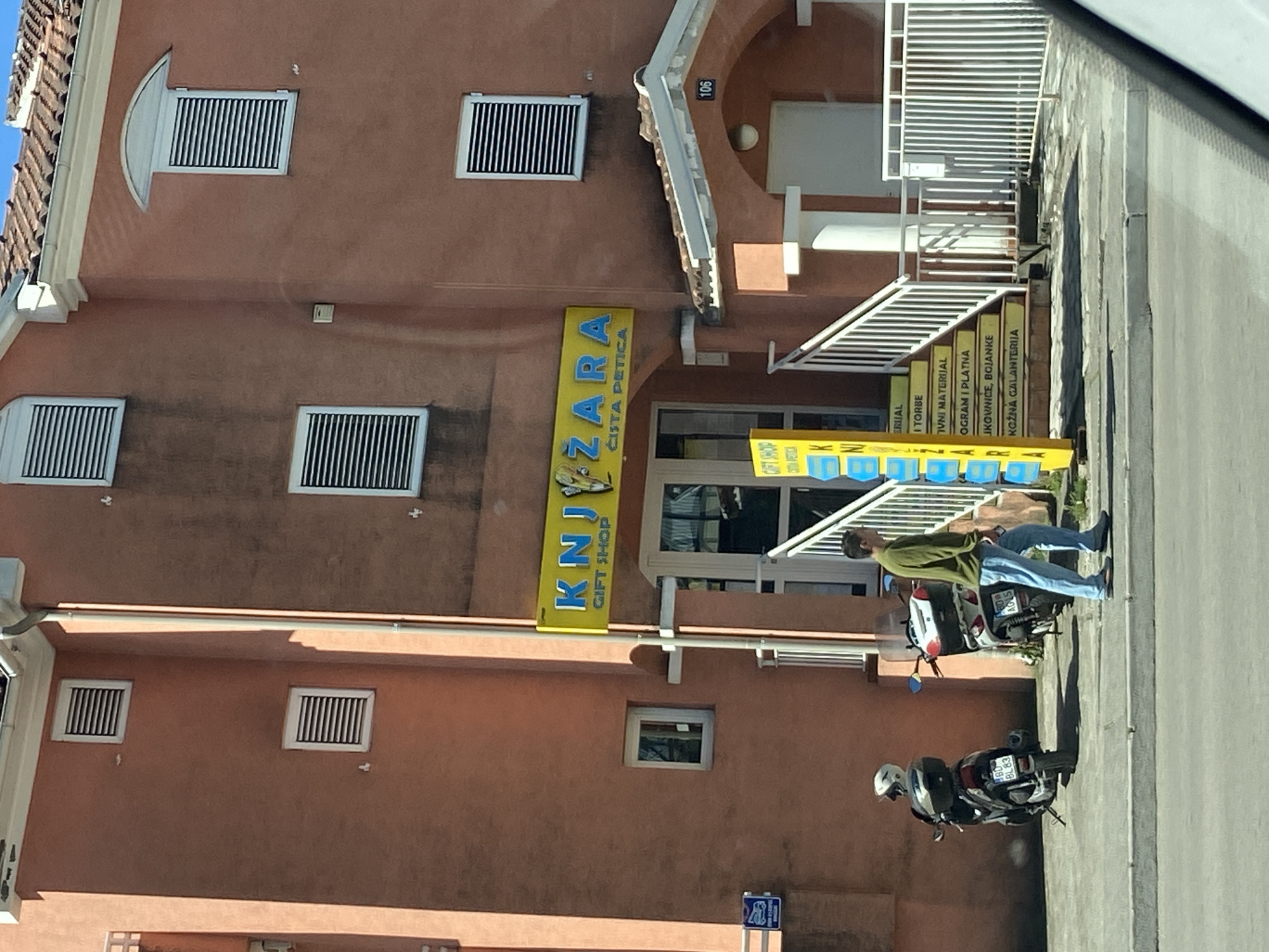 Here is a detailed description of the image:
**Scene:**
The image captures an exterior view of a multi-story building with a light orange facade. The building is identified by a yellow vertical sign labeled "KNJIŽARA GIFT SHOP ČISTA PETICA" displayed next to the entrance. The windows are rectangular and have dark horizontal slats. A white, modern-style staircase leads to an upper level of the building.
**Subjects & Objects:**
- A man is seen leaning against a parked motorcycle near the entrance of the building.
- Two motorcycles are parked along the curb. One is white and the other is black.
- The vertical sign has text in both English and what appears to be Croatian. This text includes different school material
- The number "106" is displayed in the top right corner.
**Location:**
Based on the text on the sign and other visual clues, the image is likely taken in the city of Budva, Montenegro.
