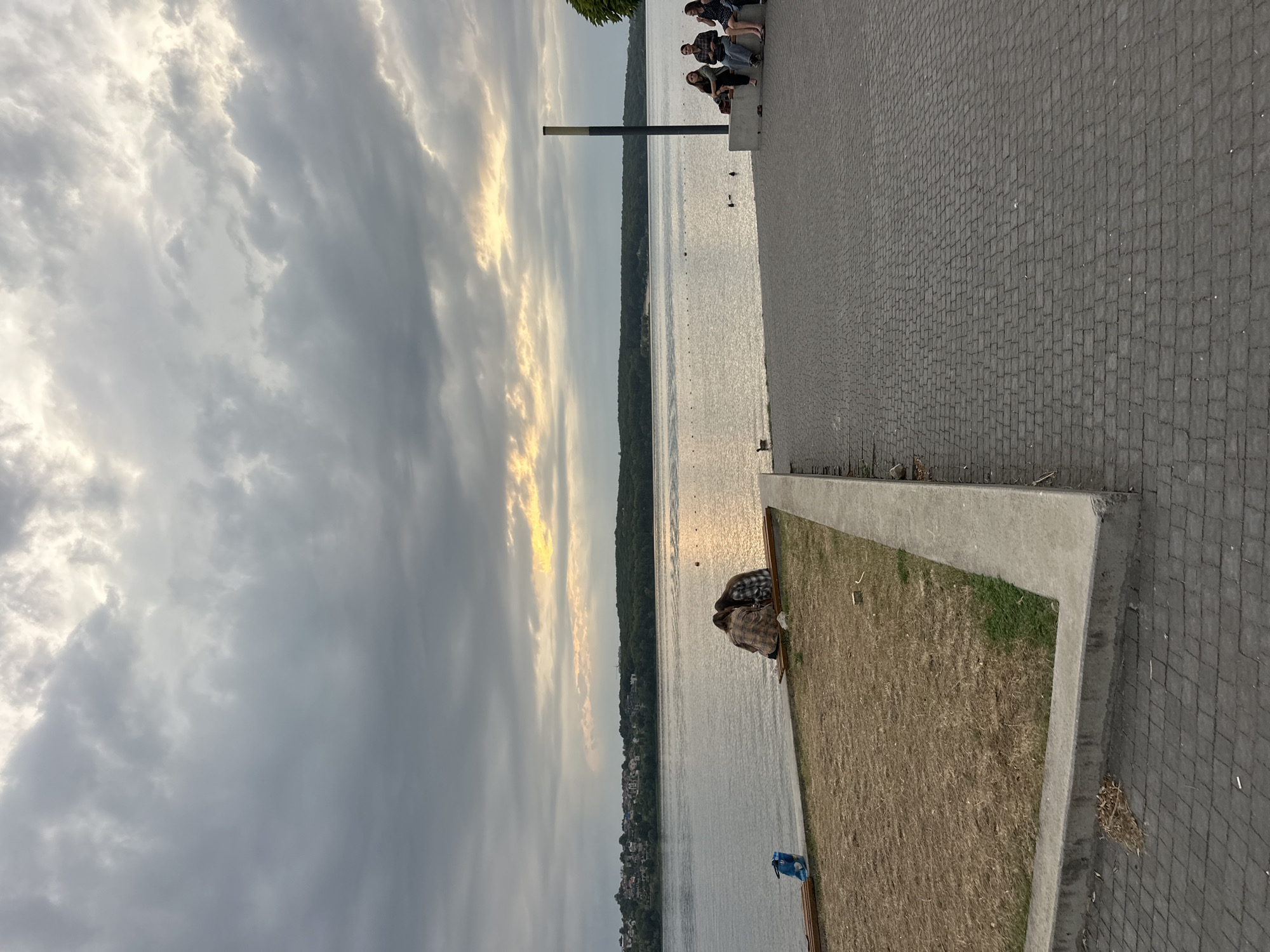 The image captures a tranquil evening scene along the Ternopil Pond in Ternopil, Ukraine. The sky is overcast with dramatic, textured clouds, but patches of sunlight break through, casting a warm glow on the water and the distant treeline. The pond itself is calm, with gentle ripples reflecting the muted light.
In the foreground, a paved walkway made of dark cobblestones stretches along the embankment. To the left, a patch of dry, brown grass is bordered by a concrete edge. Two individuals are seated close together on a wooden bench at the edge of the grass, facing the water. One is leaning their head on the other's shoulder, suggesting intimacy or comfort. A blue bag is placed on the ground near them.
Further along the walkway, a group of four people are seated on a raised concrete structure, appearing to converse or relax. A tall, thin pole with a dark top stands nearby. A goose is visible in the water to the left of this group, adding a touch of wildlife to the scene.
The opposite bank of the pond is lined with a dense forest, and beyond that, the rooftops of houses and buildings of a settlement can be seen. The overall atmosphere is peaceful and serene, a typical evening at a waterfront promenade.