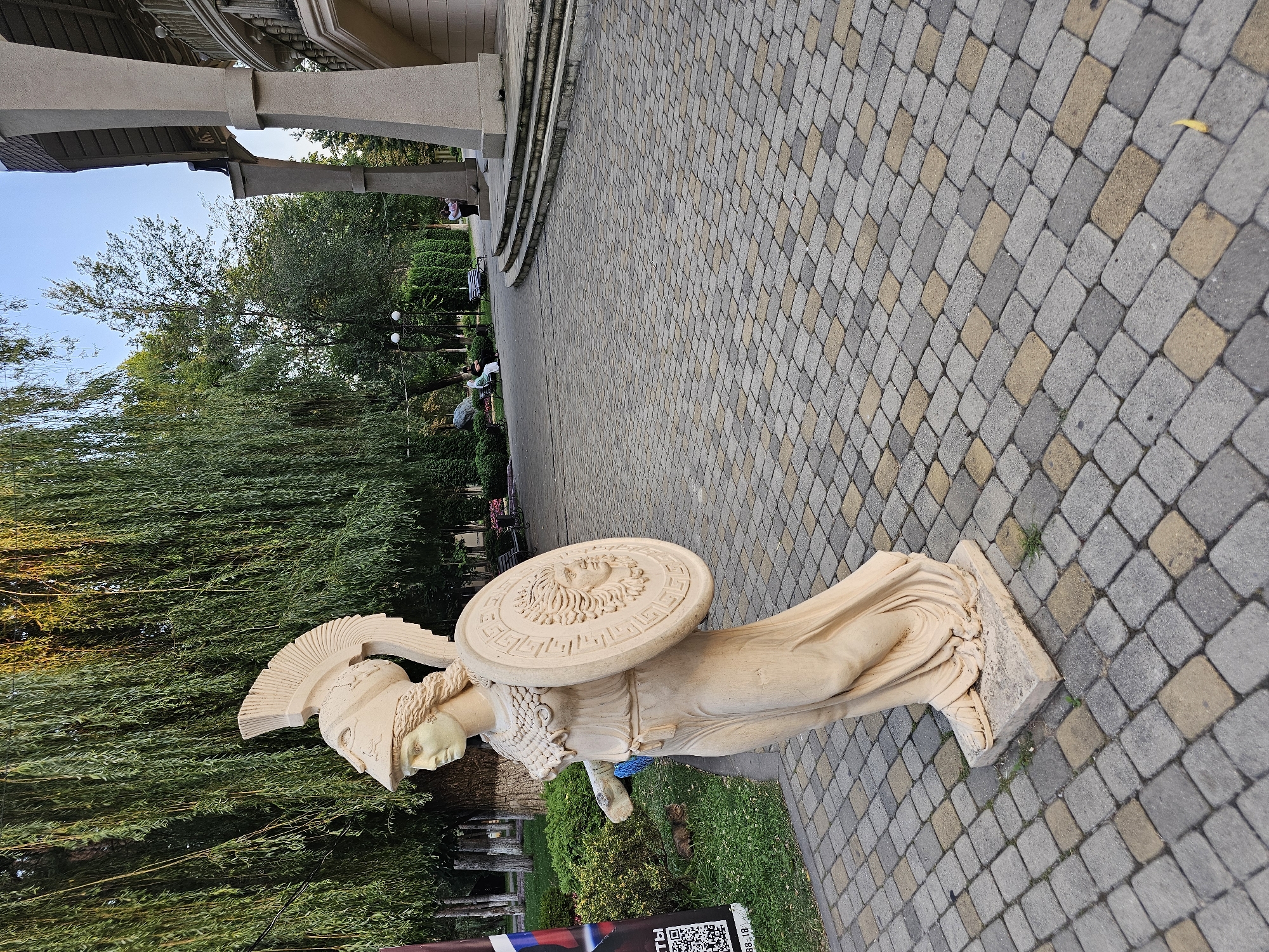 This is a full-shot, eye-level, outdoor photograph taken in Cherkessk, Russia. The primary subject is a beige stone statue of a woman dressed in ancient Greek attire, wearing a helmet and holding a shield. The statue is positioned on a paved pathway composed of interlocking gray and tan rectangular stones. To the left of the statue, there is a tall, weeping willow tree with lush green foliage, and further back, a dark-colored banner with Russian text and a QR code is visible. In the background, there is a park-like setting with more trees, benches, and a building with a grand staircase supported by large, cylindrical columns. A few people are casually seated on benches in the distance. The lighting suggests it is either early morning or late afternoon, with sunlight casting a warm glow on the statue and tree.