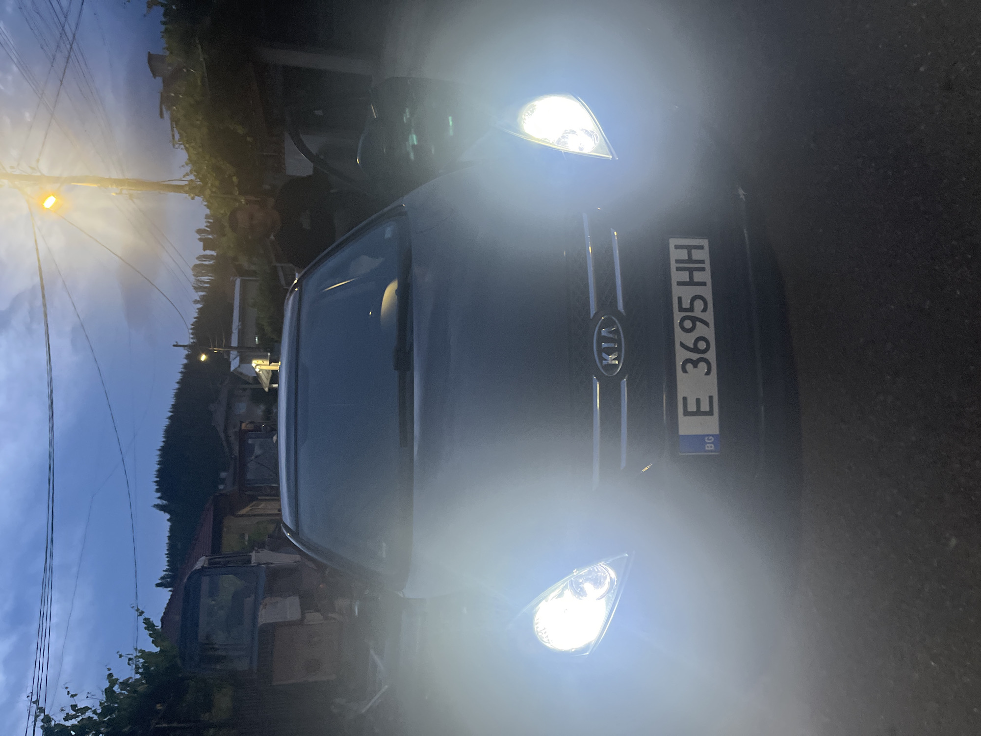 The image captures a dark-colored Kia car head-on at what appears to be dusk or nighttime in Pokrovnik, Bulgaria. The car's bright headlights are illuminated, casting significant light spill and lens flares across the foreground. The Kia logo is clearly visible on the grille.
Behind the windshield, a person's upper body and head are partially visible, suggesting they are seated in the driver's seat. The license plate is clearly readable as "E 3695 HH," with a "BG" country identifier indicating Bulgaria.
In the background, under a darkening sky, a street light is on, emitting a warm, orange glow and highlighting power lines crisscrossing the frame. Further back, densely forested hills or mountains are visible, silhouetted against the twilight sky. Buildings and structures, including what appears to be an older excavator or piece of construction equipment, are discernible through the dim light, nestled among trees and foliage. The overall setting appears to be a rural or semi-rural street or driveway.