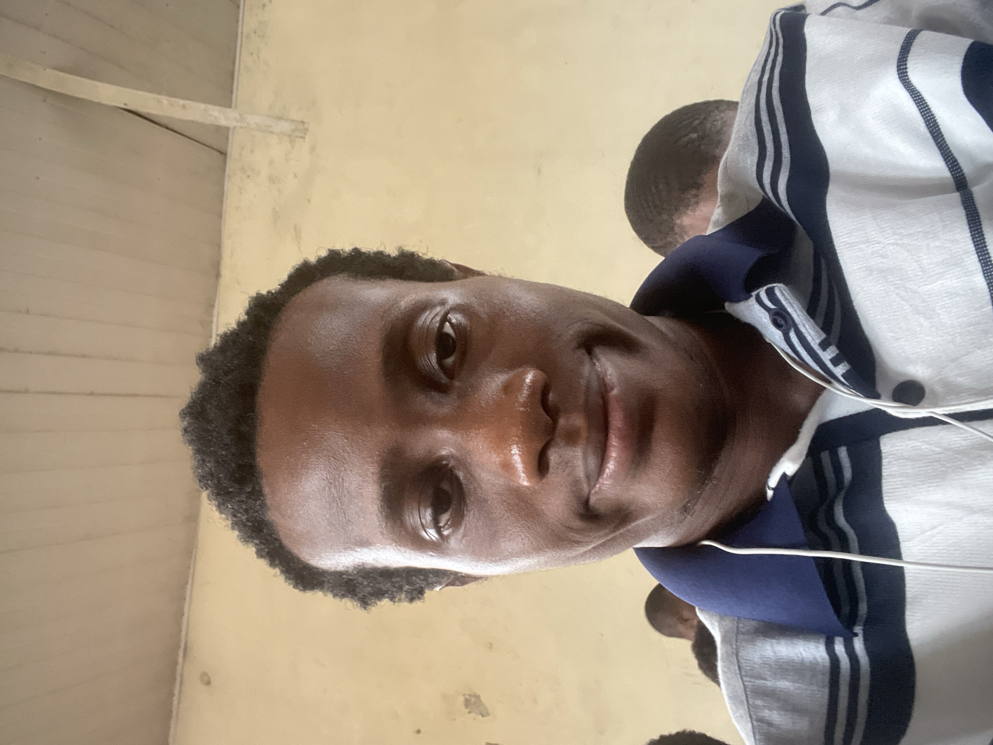 In the city of Ihiagwa, Nigeria, a young Black man, likely in his early twenties, is captured in a close-up, front-facing selfie. He has a dark complexion, short, textured black hair, and appears to be wearing a striped polo shirt. A white earphone cord hangs from his shirt collar, suggesting he might be listening to something. The background is nondescript, with a light-colored, possibly painted wall and a wooden ceiling, indicating an indoor setting. Hints of other individuals can be seen in the blurred background, suggesting the presence of others in the vicinity. The lighting is even, suggesting the photo was taken in daylight or under artificial light. The overall impression is of a casual, everyday moment.