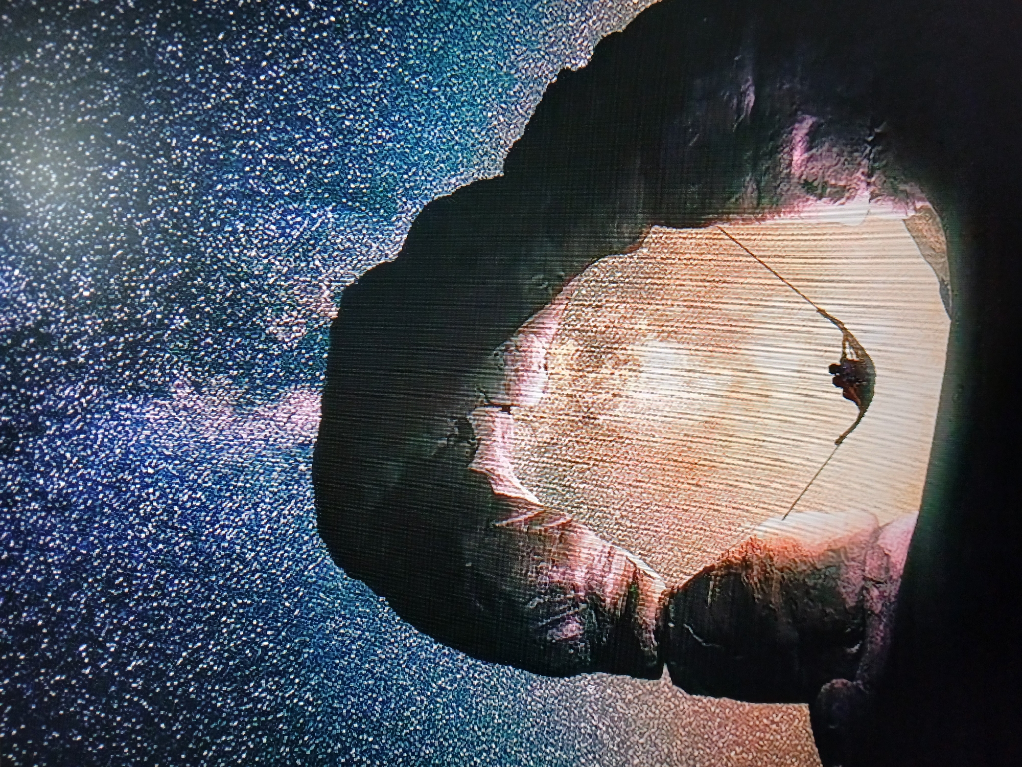 The media file depicts a dark, rocky cavern opening to a bright, starry expanse. The cavern's interior is lit by an unseen light source, casting a warm, orange glow on the rough, textured surfaces of the rock formations. A thin rope extends from the right side of the frame, disappearing into the darkness and suggesting a climber or explorer suspended within the cavern. The exterior is a deep blue, densely populated with small, bright white specks representing stars. The overall impression is one of awe and exploration, with a sense of a vast, unknown environment. The texture of the image suggests it may be a digital rendering or a heavily processed photograph. No text is visible.