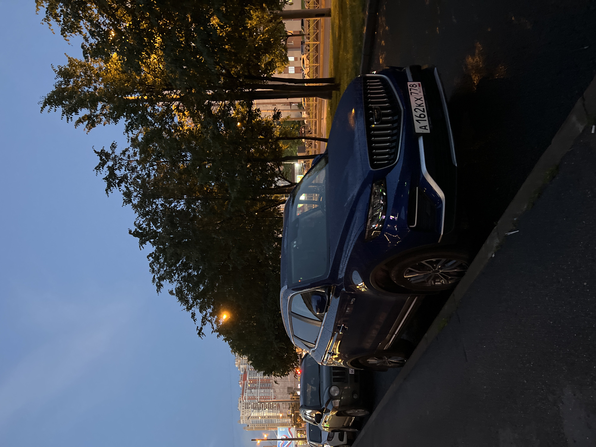 The image shows a blue SUV parked on the side of a road at dusk. The sky is a deep blue, transitioning from light to dark, suggesting the end of the day. Trees with dense foliage line the road, their leaves illuminated by artificial lights, creating a warm glow. In the background, a cityscape with buildings of various heights is visible, some brightly lit. The road surface appears wet, indicating recent rain or dew, and reflects the surrounding lights.
The car, a modern SUV, is the primary object in the foreground. Its license plate, partially visible, reads "A 162KX 778". The car is positioned at an angle, with its front grille and headlights clearly discernible. The overall scene is one of urban twilight, with a blend of natural and artificial elements. The atmosphere seems calm and quiet, typical of the evening in a city. The lighting suggests it is either early morning or late evening. There are no people visible in the frame. The setting appears to be a street in a Russian city, given the format of the license plate.