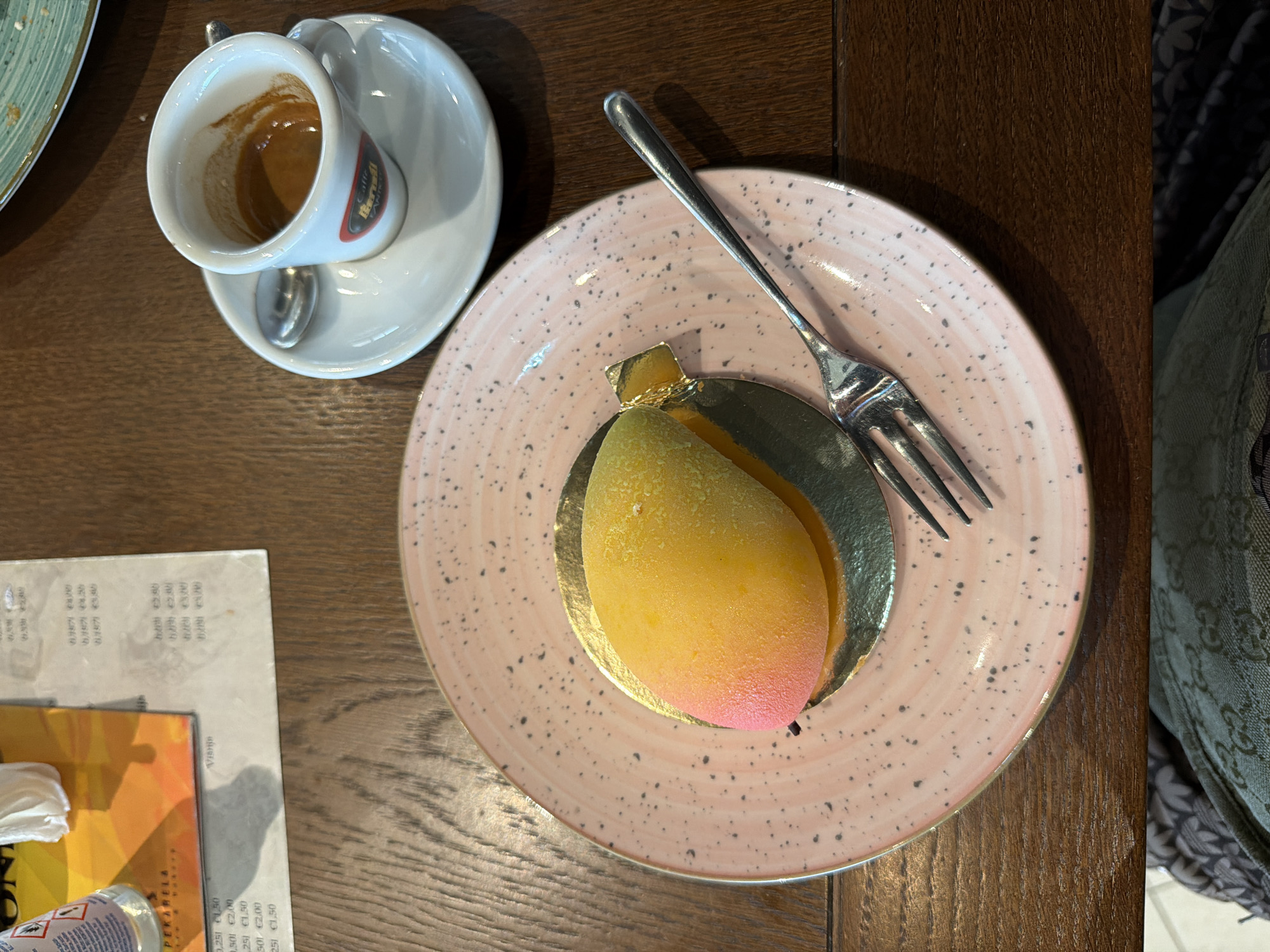 The image shows a wooden table in what is likely a café in Budva, Montenegro. On the table, from left to right, are a partially visible plate, a white espresso cup and saucer with a spoon, a pink plate containing a mango-shaped dessert with a silver fork, and a menu/bill. To the right of the plate is a patterned item, likely clothing. The dessert has a golden base and a gradient color going from yellow to pink.
--- OCR End ---