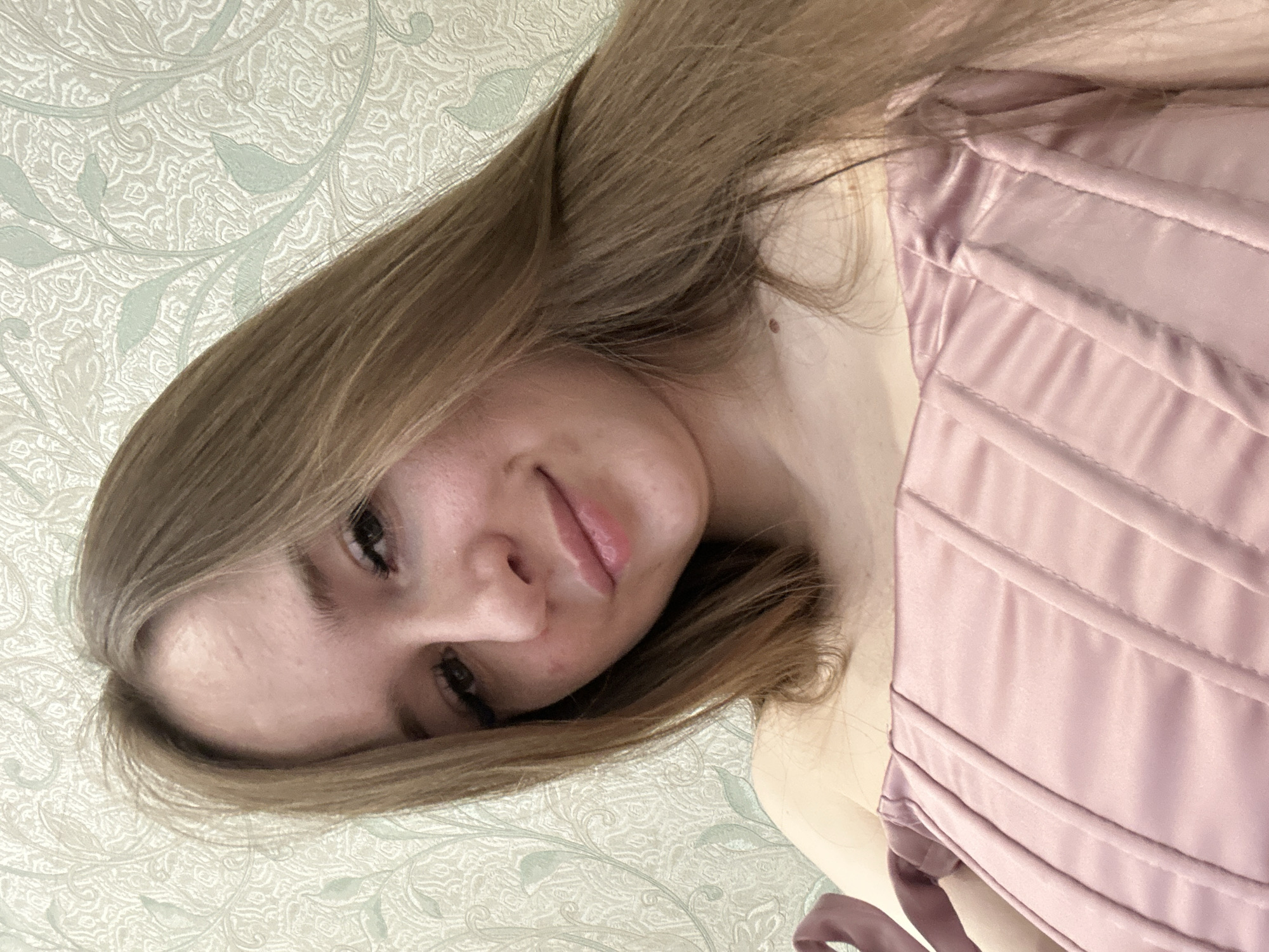 A close-up, slightly rotated image features a young woman with light brown hair. She is wearing a light pink corset-style top. Her face is turned towards the camera, with a slight smile. The background is a light-colored patterned wall, with a floral motif.