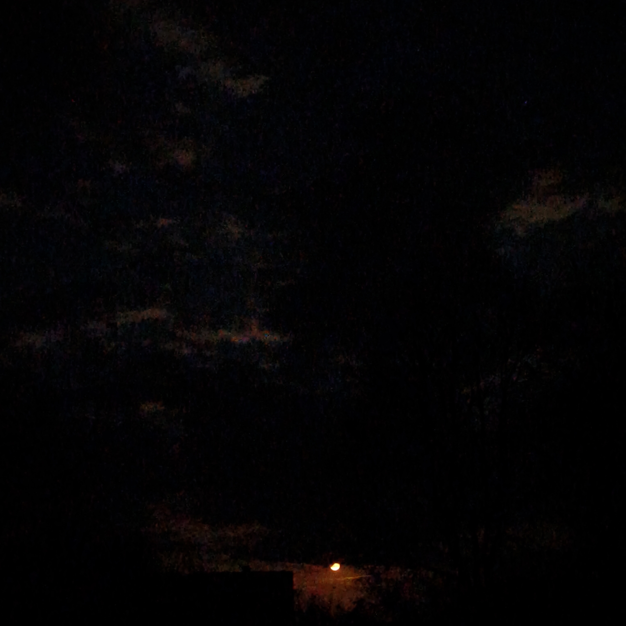 The image depicts a night scene in Парэчча, Belarus. The sky is dark with indistinct clouds. In the lower part of the image, there is a silhouette of a building or structure. To the right, a street light emits a bright orange glow. The surrounding environment is dark and indistinct, possibly containing vegetation.
