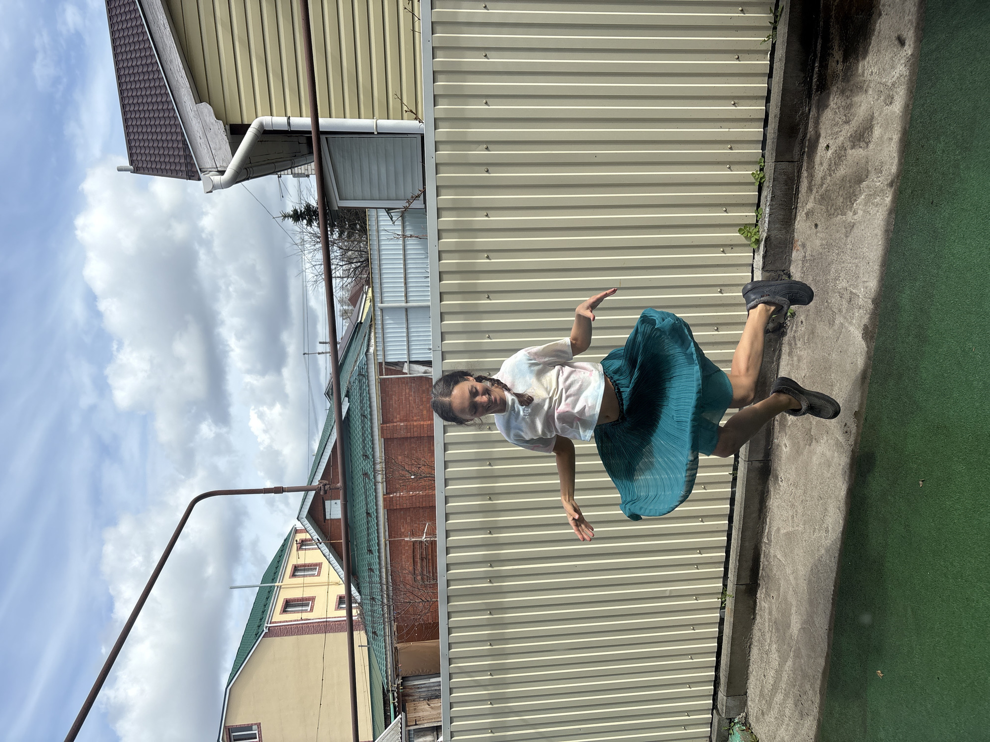 A woman is suspended in the air in front of a corrugated metal fence. She is wearing a white shirt, a teal pleated skirt, and dark-colored shoes. She has her arms outstretched, palms facing forward. The location appears to be a backyard or alleyway in Ulyanovsk, Russia, based on the architectural styles of the surrounding buildings. The buildings are a mix of yellow and brick, with green and brown roofs. A utility pole is visible on the left side of the frame. The ground is a mix of concrete and what appears to be green artificial turf. The sky is visible behind the buildings, with a mix of clouds and blue sky.