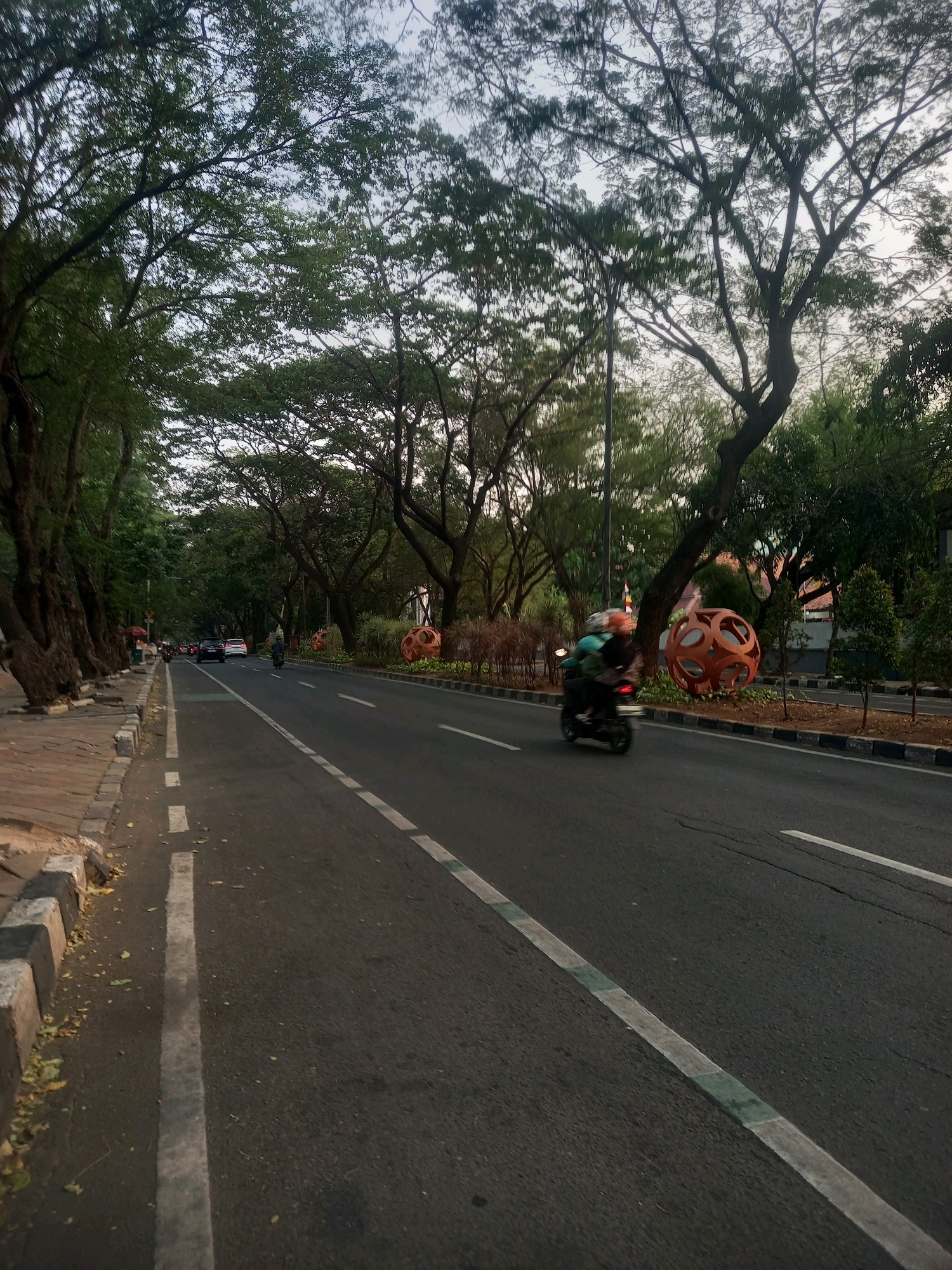 The image depicts a street scene in Tangerang, Indonesia, characterized by a tree-lined avenue with a paved road and a sidewalk. The road has two lanes of traffic with white and green lane markings. A motorcycle is in motion on the right lane, with its rider blurred due to the speed. Several cars are also visible further down the road. The left side of the road features a sidewalk with stone curbing, and large trees with dense foliage line the street. In the median of the road, there are decorative orange spherical sculptures. The overall lighting suggests it is either early morning or late afternoon.