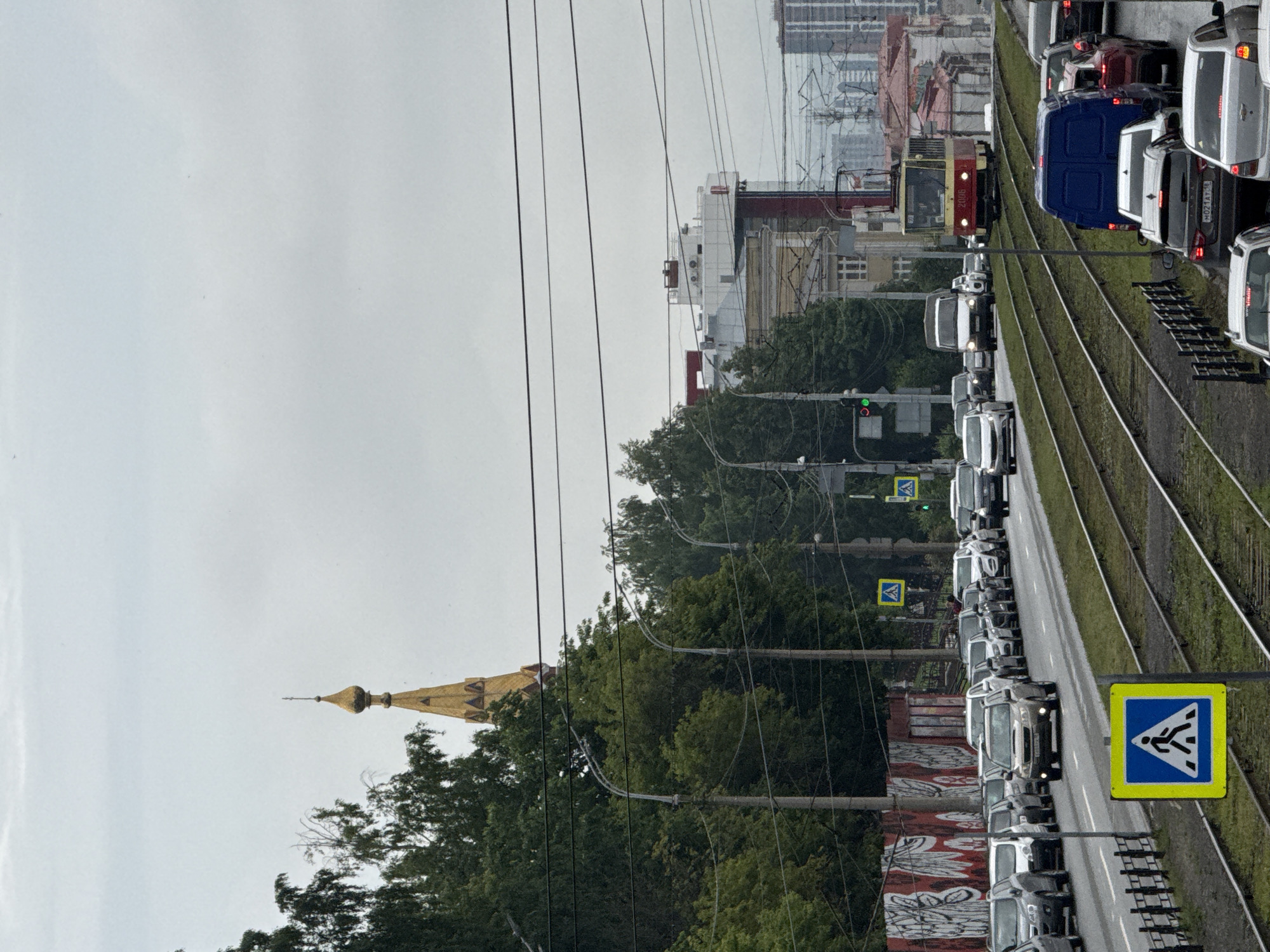 The image shows a street scene in Izhevsk, Russia. The foreground features tram tracks and a pedestrian crossing sign. A traffic jam of cars and a blue van is visible on the right side of the tram tracks. Further down the street, a red and yellow tram is traveling on the tracks. On the left side of the street, there are more cars, traffic lights, and trees. A golden dome of a church is visible above the trees in the background, and several buildings line the street. The sky is overcast.