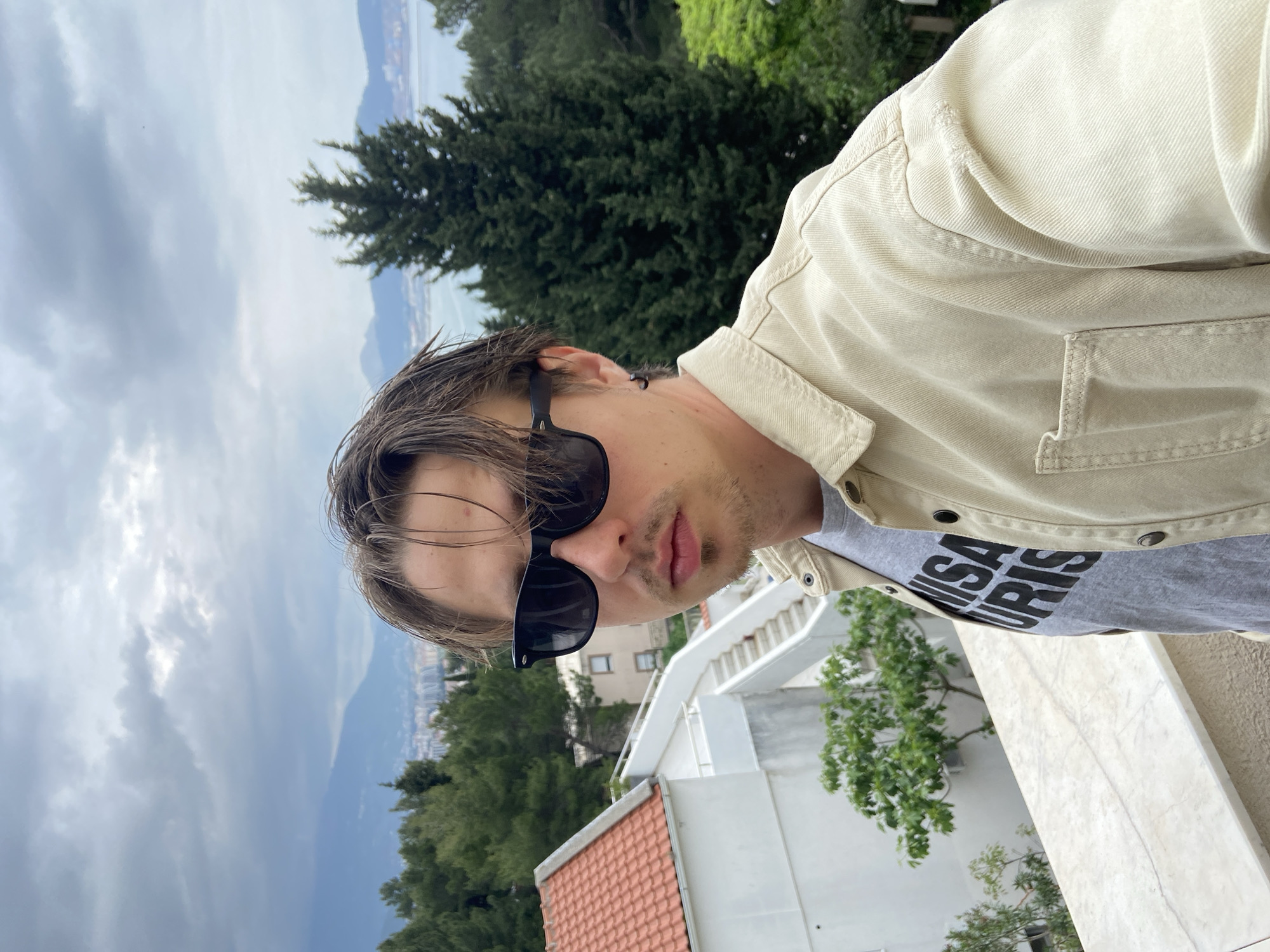 A man wearing sunglasses and a tan jacket stands on a balcony in Šušanj, Montenegro. The background includes a cloudy sky, distant mountains, the Adriatic Sea, trees, and buildings. The man wears a gray T-shirt with black lettering. The balcony has a white marble railing. A building with a red tile roof and white exterior is visible below the balcony.