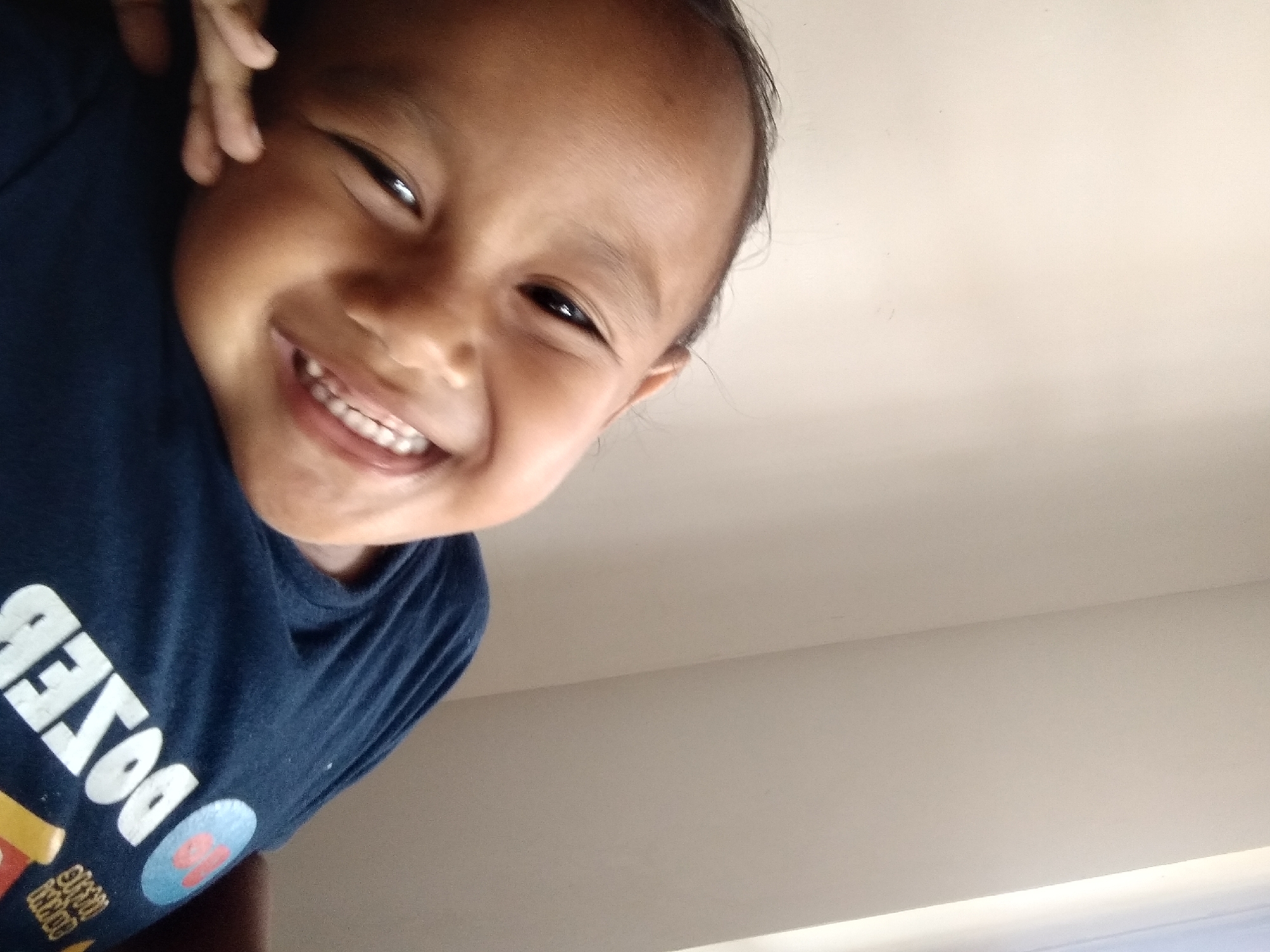 A close-up, indoor, portrait photograph shows a young child smiling broadly, their head tilted to the left. The child is wearing a dark blue t-shirt with white lettering that appears to say "DOSEB" in large, bold font, along with some other graphic elements. The background consists of a plain, light-colored wall, with a sliver of bright light visible on the right, suggesting a window or light source. The lighting is soft and natural. The image has a casual, personal feel, indicative of a personal photograph. The location is identified as Pandak, Indonesia.