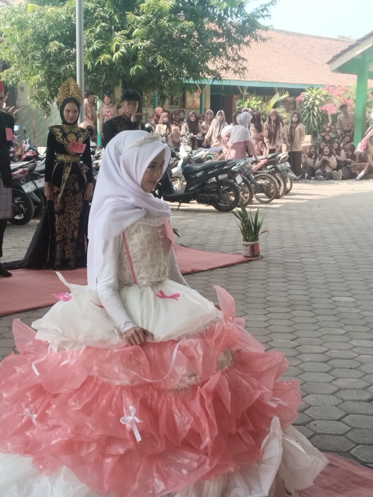 In Pakulaut, Indonesia, a young woman wearing a white hijab and a layered, voluminous dress made of pink and white ruffles sits on a pink mat in front of a group of people. The dress has a corseted top with a floral pattern and pink ribbon accents. She is looking towards her right. To her left, another young woman in a black and gold traditional outfit with a gold headdress stands with a red and white tag labeled "012" pinned to her chest. Behind them, a gathering of individuals, many in uniforms, are visible, along with several parked motorcycles. The scene appears to be an outdoor event taking place on a paved area with trees and buildings in the background.