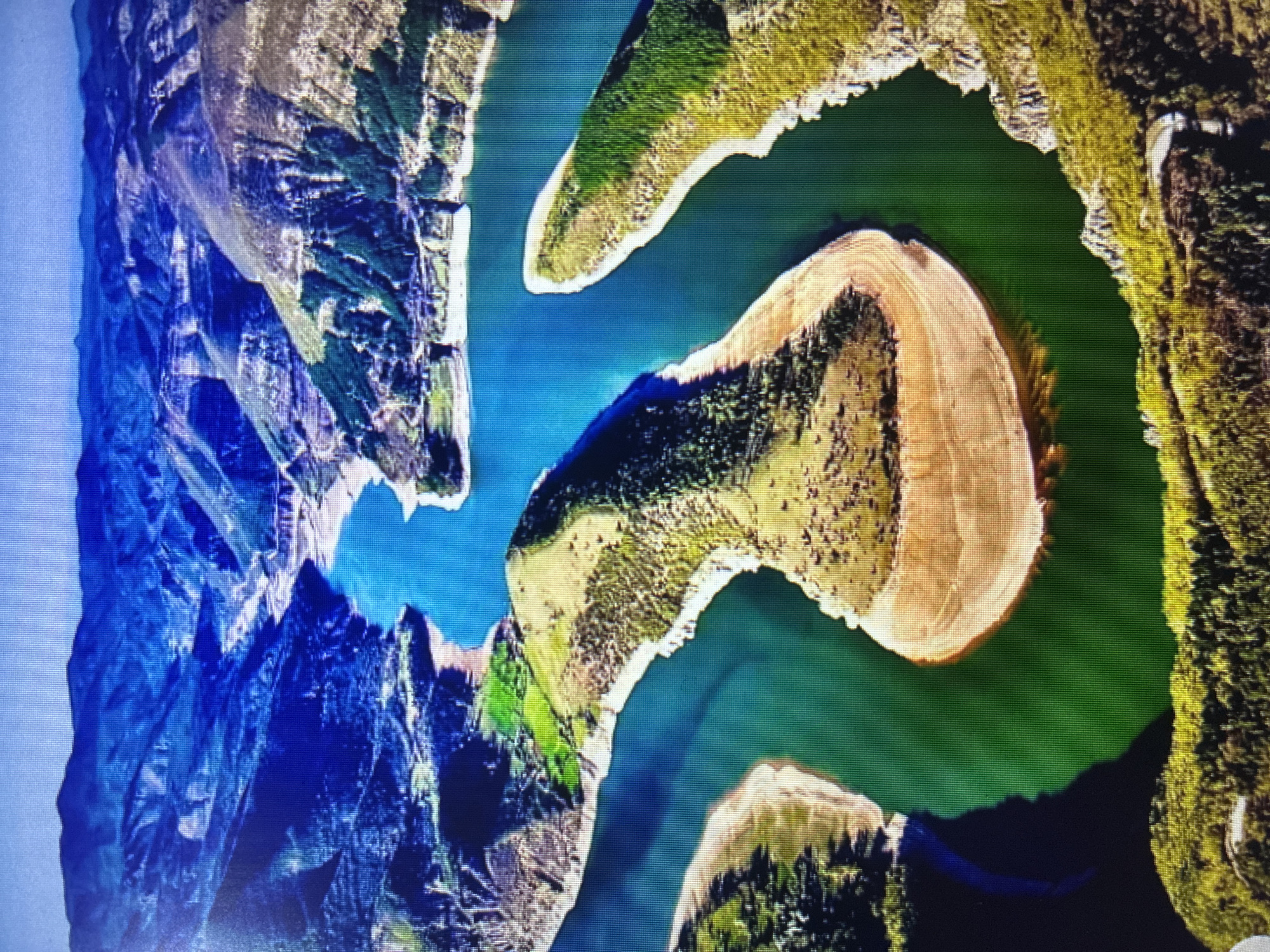 This is an aerial photograph depicting a winding body of water, likely a river or reservoir, surrounded by mountainous and hilly terrain. The image, appearing to be a photograph of a screen due to visible pixelation, is rotated 90 degrees counter-clockwise, showing the water stretching from the top-left to the bottom-right.
The water exhibits a striking range of colors, from deep emerald green in some sections to a lighter, clearer blue-green, suggesting varying depths or mineral content. Several prominent landmasses jut into the water, forming distinct meanders. One large, U-shaped peninsula, primarily light brown with some scattered vegetation, is particularly notable in the center of the frame. The surrounding land is rugged, characterized by steep, rocky cliffs and slopes covered with dense dark green forests and lighter green vegetation. Blue and grey tones define the distant mountainous peaks.
The scene depicts a natural landscape, with no visible signs of human activity or structures, emphasizing its remote and pristine character. The lighting indicates it is daytime under clear weather conditions, casting subtle shadows that highlight the topography. This landscape is consistent with the mountainous regions and reservoirs found near Asenovgrad, Bulgaria. No text is visible in the image.