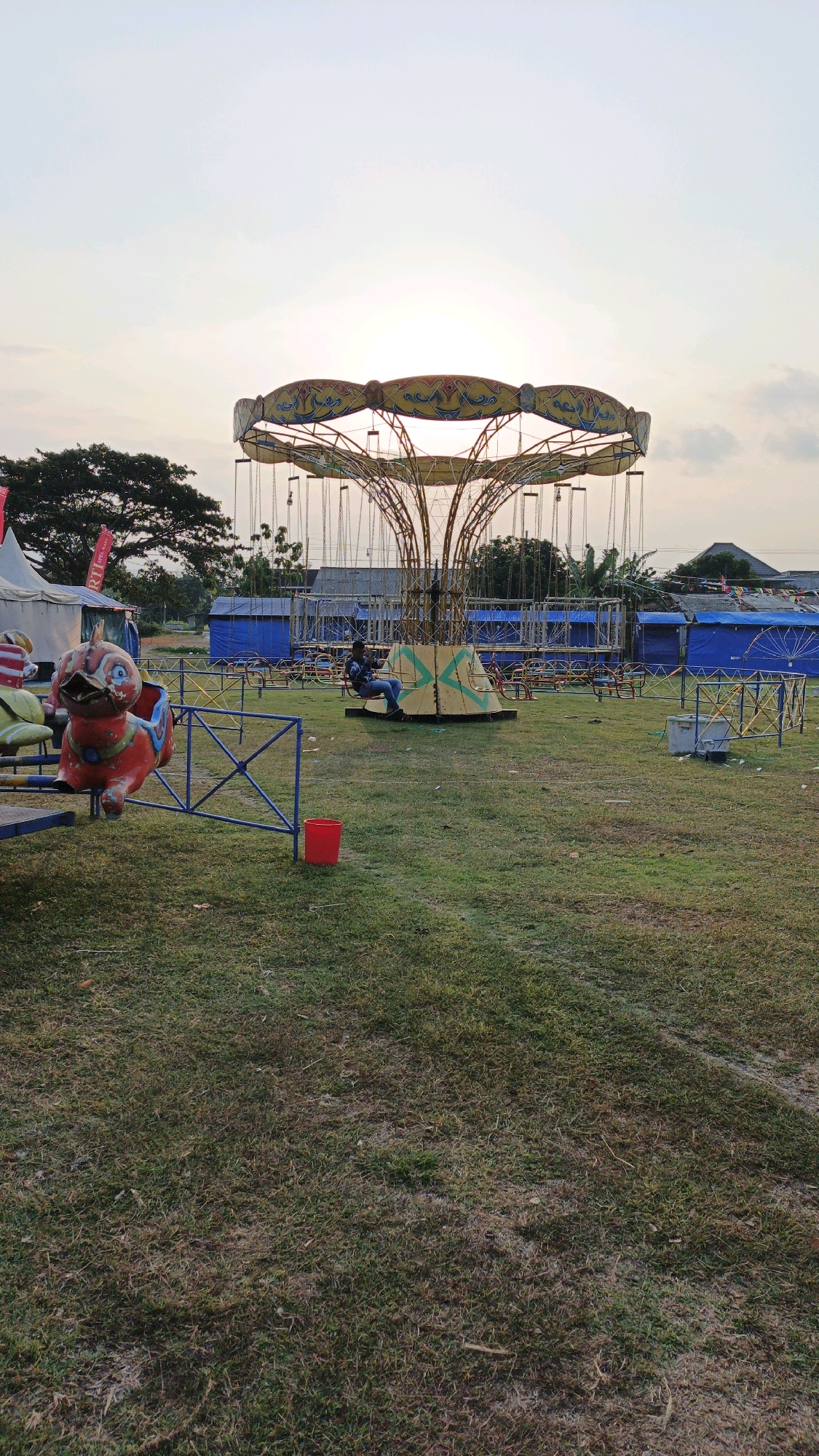 In Boyolangu, Indonesia, at dusk, a carousel ride stands in a grassy field. The ride, adorned with yellow and red, is empty except for a single person sitting in one of the swinging chairs. The sun is setting, casting a warm glow over the scene. In the foreground, a red bucket and a blue fence are visible. In the background, tents, blue structures, and trees complete the outdoor amusement park setting.