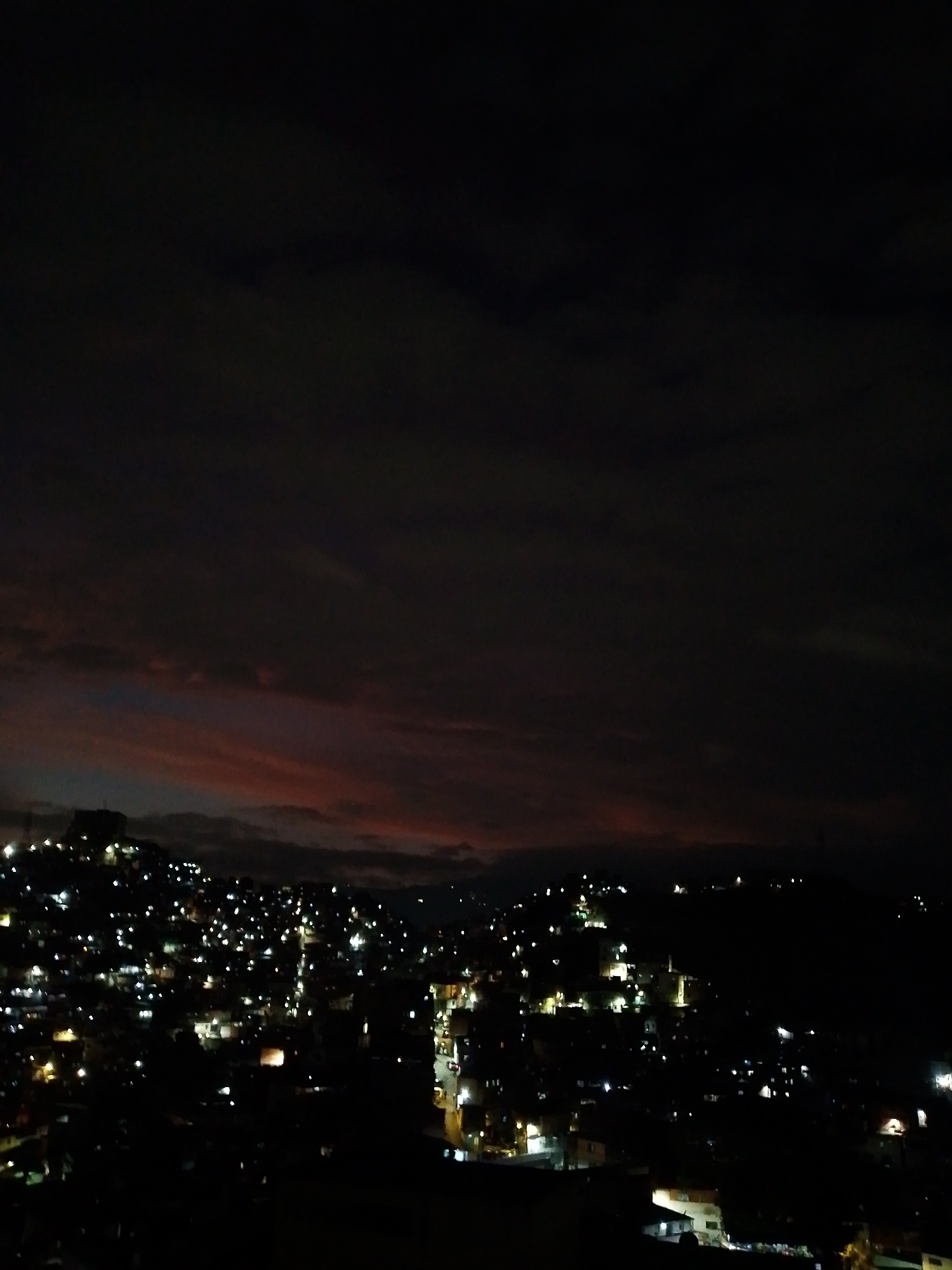 The image shows a nighttime view of Caracas, Venezuela. The city is visible in the foreground and middle ground, with many lights from buildings and streets illuminating the landscape. The sky is dark, with some clouds visible near the horizon, possibly reflecting the city lights. The overall scene is dark, emphasizing the urban setting and nighttime atmosphere.