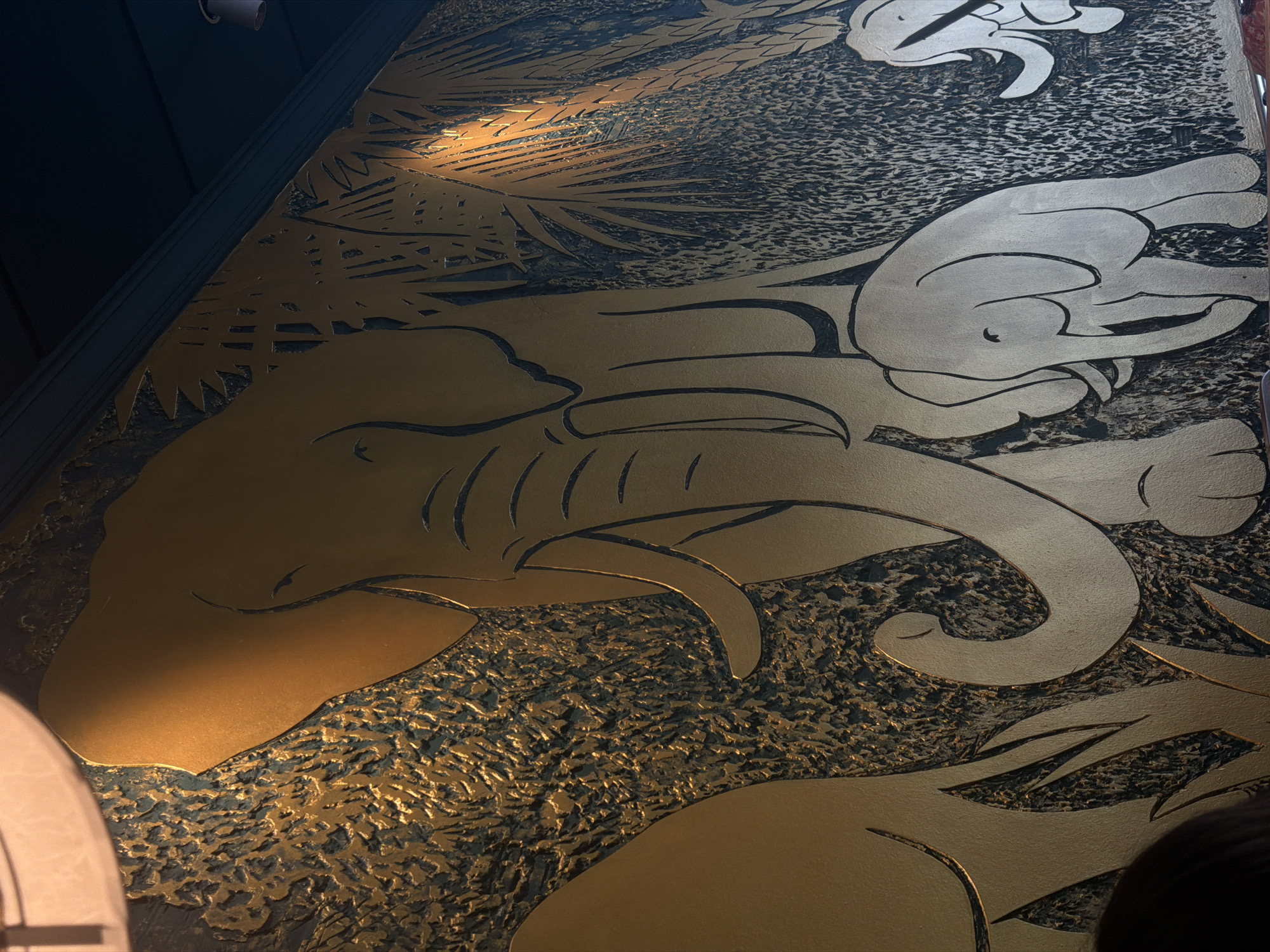 This media file captures a close-up view of a wall or panel adorned with a decorative relief sculpture. The artwork features stylized depictions of elephants and palm fronds, rendered in a metallic gold against a textured, dark background. The larger elephant in the foreground is intricately detailed, with a prominent trunk and tusks. Smaller elephant forms are visible above and to the right, also rendered in a simplified, almost abstract style. The palm fronds are sharp and angular, adding a graphic quality to the design. The surface of the wall has a rough, almost chiseled texture in the dark areas, contrasting with the smooth, polished finish of the gold elements. A warm, directional light source illuminates the scene from the upper left, casting highlights and shadows that emphasize the three-dimensionality of the relief. The lighting creates a dramatic effect, drawing attention to the curves and contours of the sculpted forms. There are no visible people or text in the image. The overall impression is one of opulence and artistic craftsmanship, possibly suggesting an interior decorative element in a restaurant, lobby, or art installation. The style evokes a sense of the exotic or tropical.
