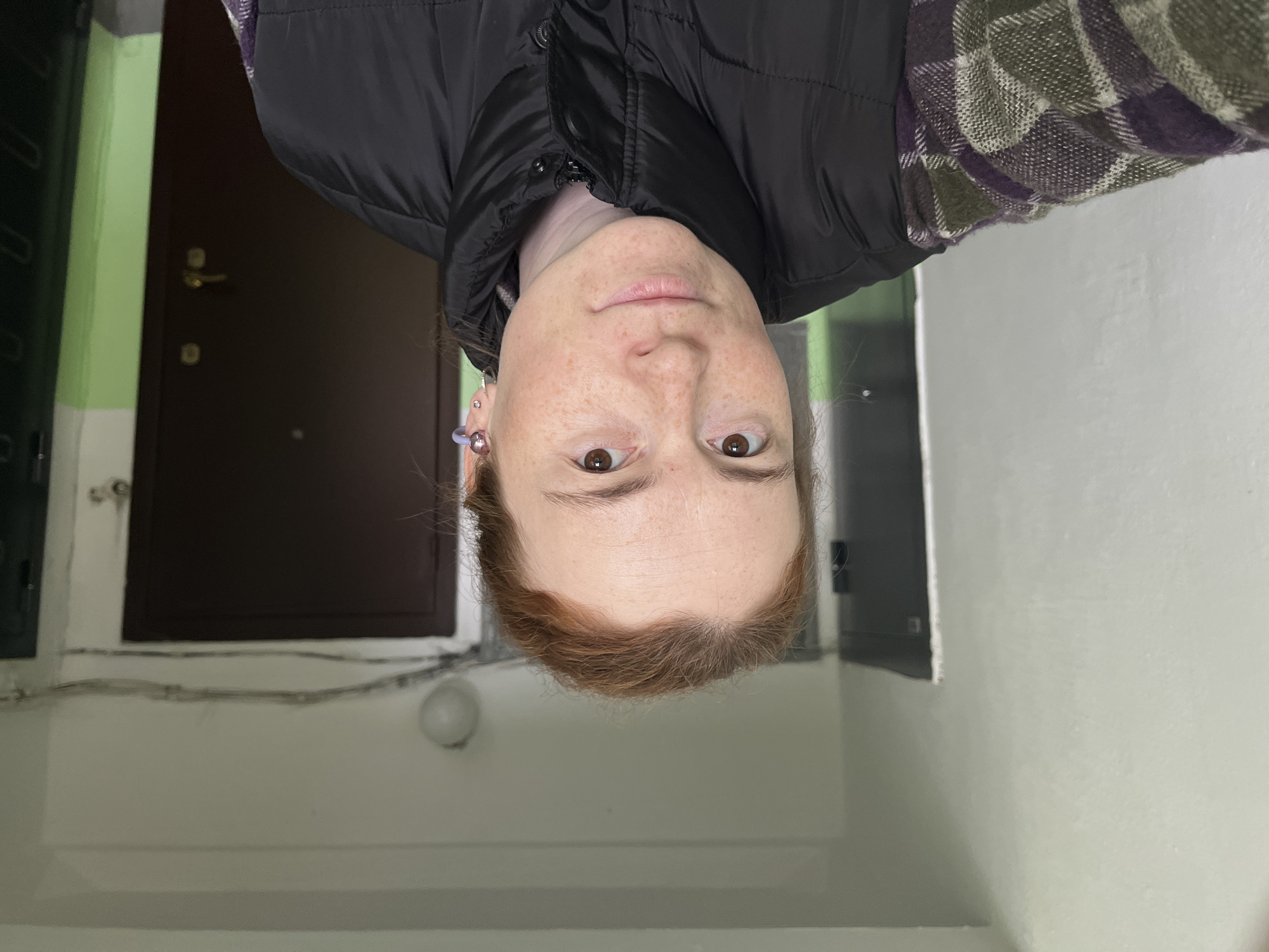 A self-portrait captures a person, likely female, looking directly at the camera in an indoor setting in Chaikovsky, Russia. The individual has fair skin with noticeable freckles, reddish-brown hair, and brown eyes. Her expression is neutral to slightly serene, with her lips closed. She is wearing a black puffer vest over a plaid shirt with a pattern of green, purple, and white squares. Multiple ear piercings are visible on her left ear, including a prominent light purple hoop or cuff and several silver-toned rings.
The background shows what appears to be a hallway or entryway. To the viewer's left, a dark brown door with a gold-colored doorknob and a silver lock is visible, flanked by a light green wall section. The lower part of the wall is painted white, with a round, frosted white light fixture affixed to it. Wires are routed horizontally along the top of the white wall. The overall lighting suggests an interior space, likely under artificial illumination. No discernible text is visible in the image.