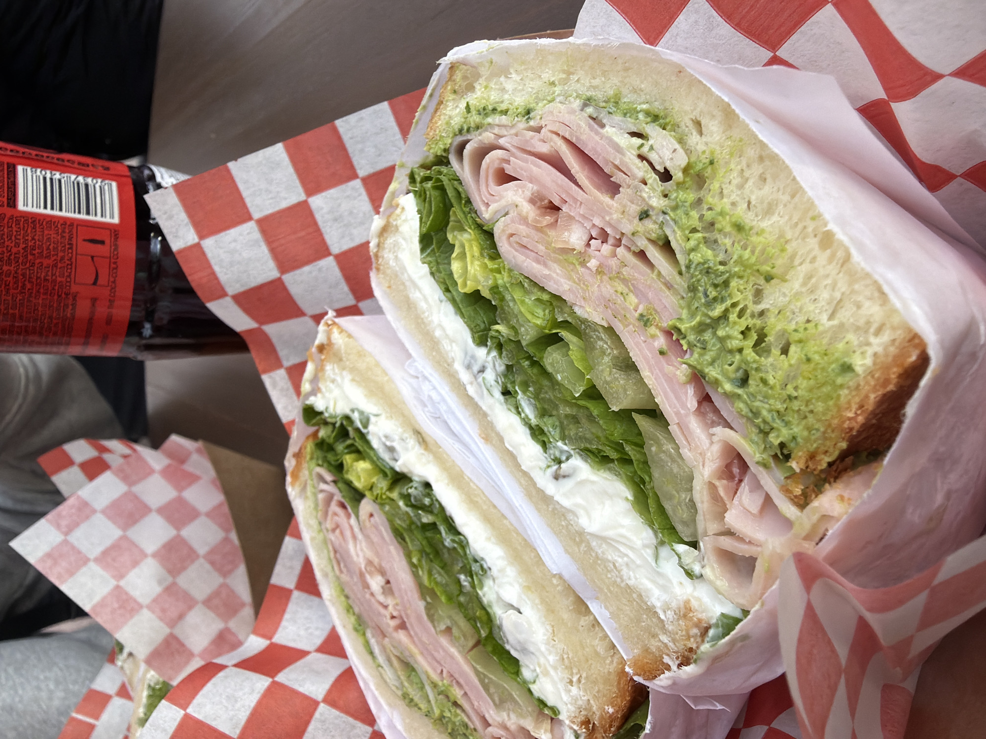 The image depicts a halved sandwich resting on a red and white checkered paper. The sandwich appears to be comprised of sliced bread, lettuce, ham, a white creamy spread, and a green spread. A bottle, possibly of Coca-Cola, is partially visible to the left of the sandwich. The location is Saint Petersburg, Russia.